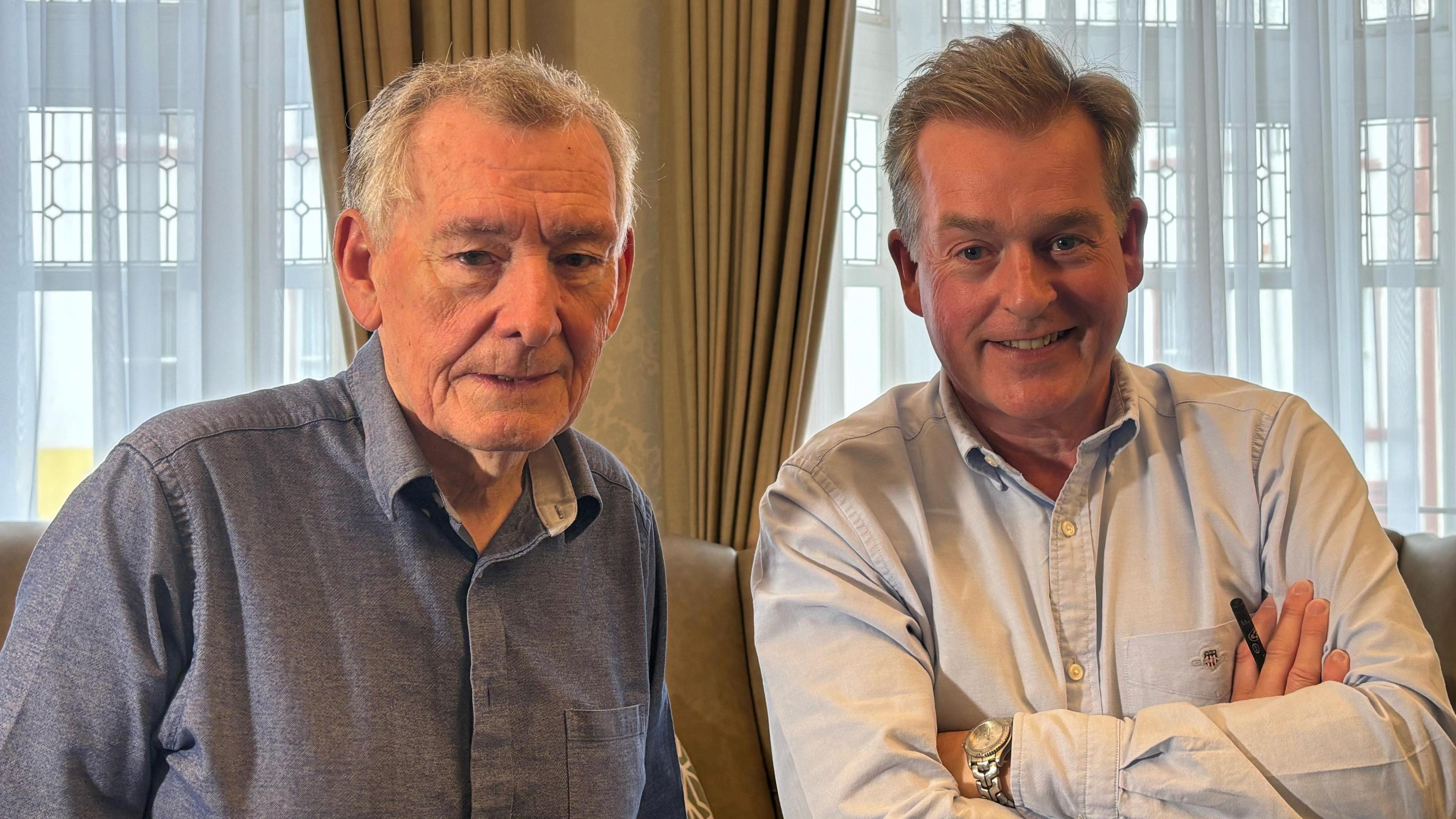 two men looking at the camera in a room with high windows and big curtains