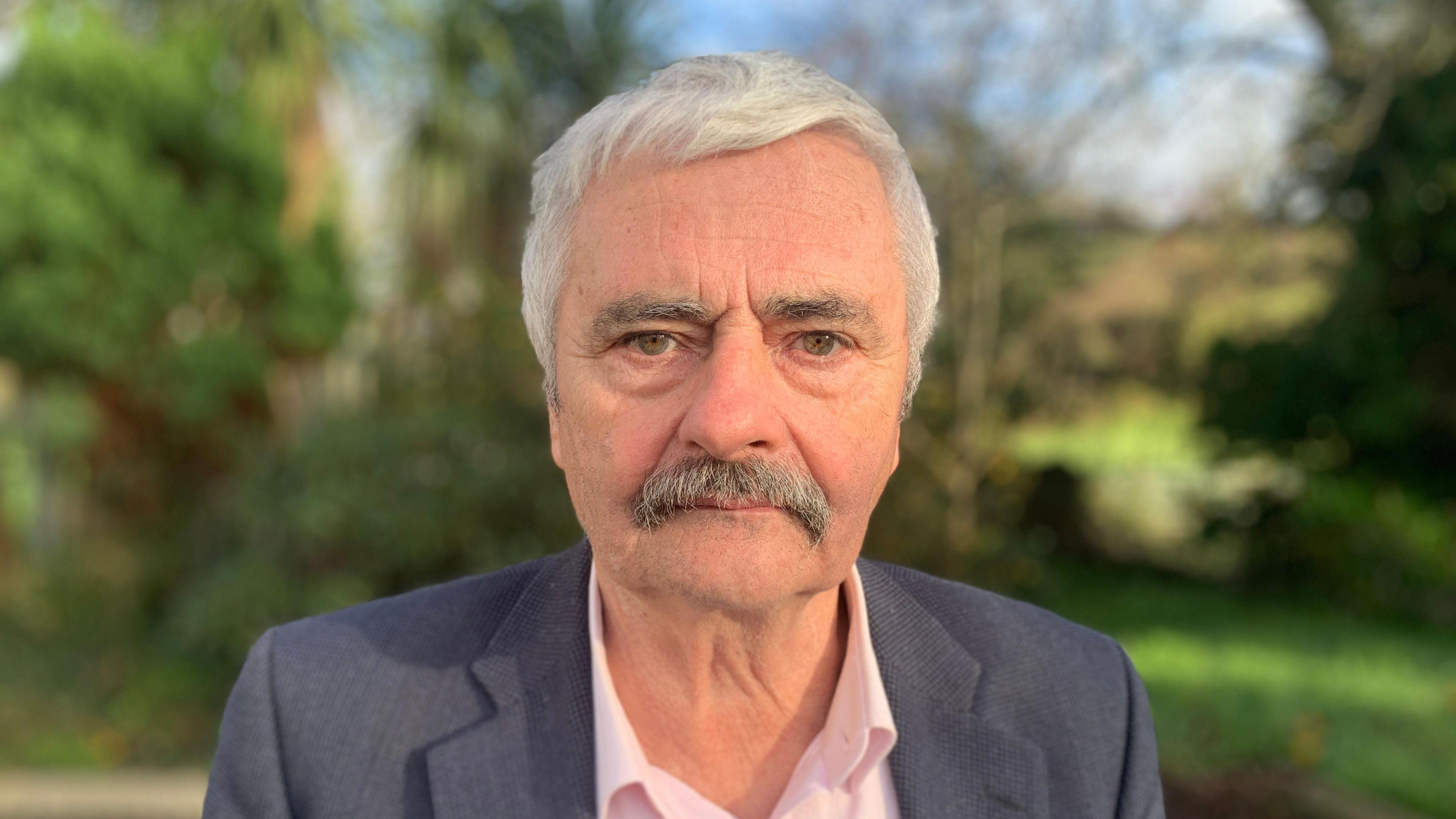 Michael Gallagher. He has white hair with grey moustache, wearing a dark grey blazer and pink shirt. Trees are blurred in the background.