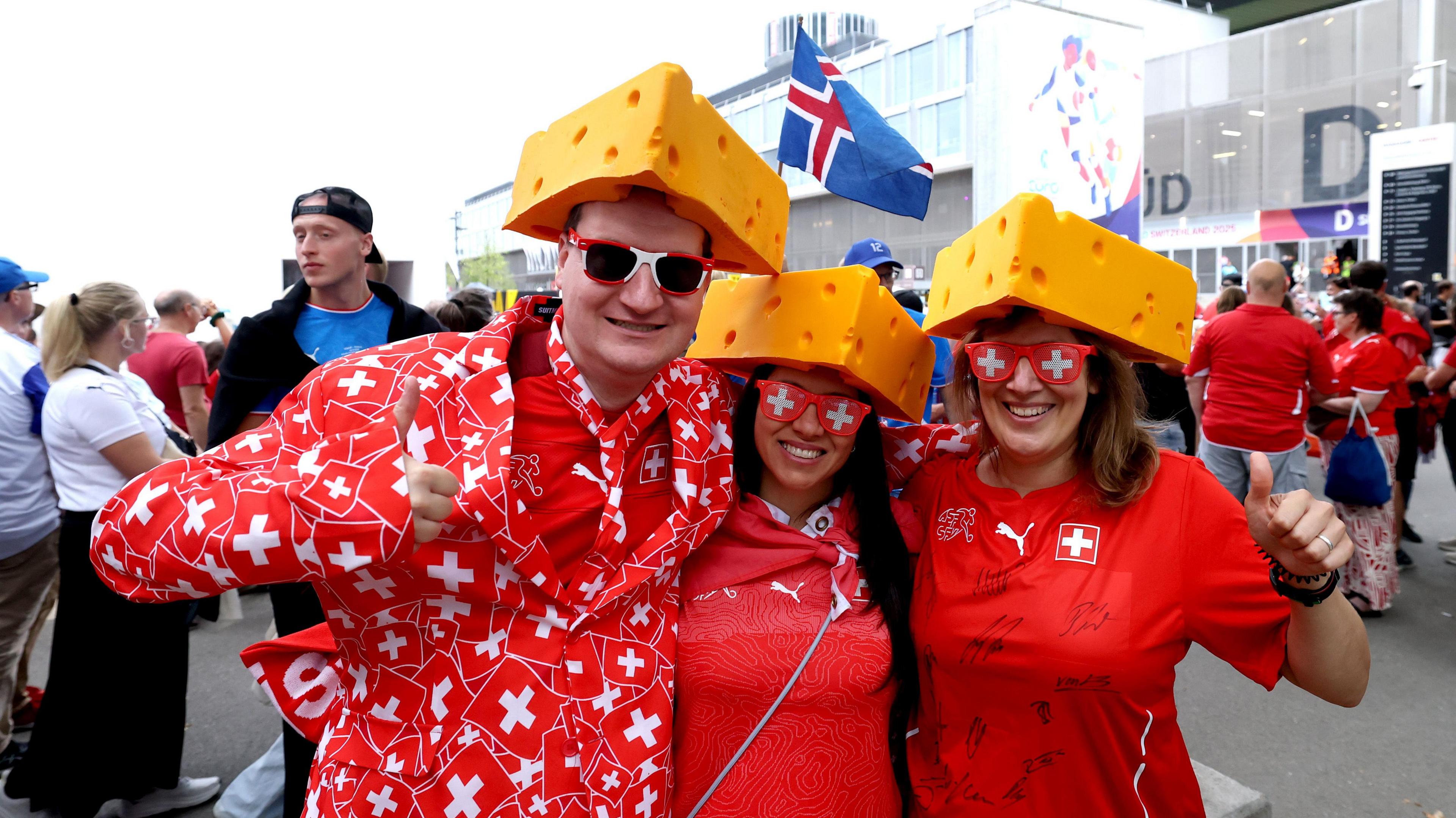 Three Switzerland fans at Euro 2025 standing together wearing read and white outfits and glasses, and yellow triangular cheese hats