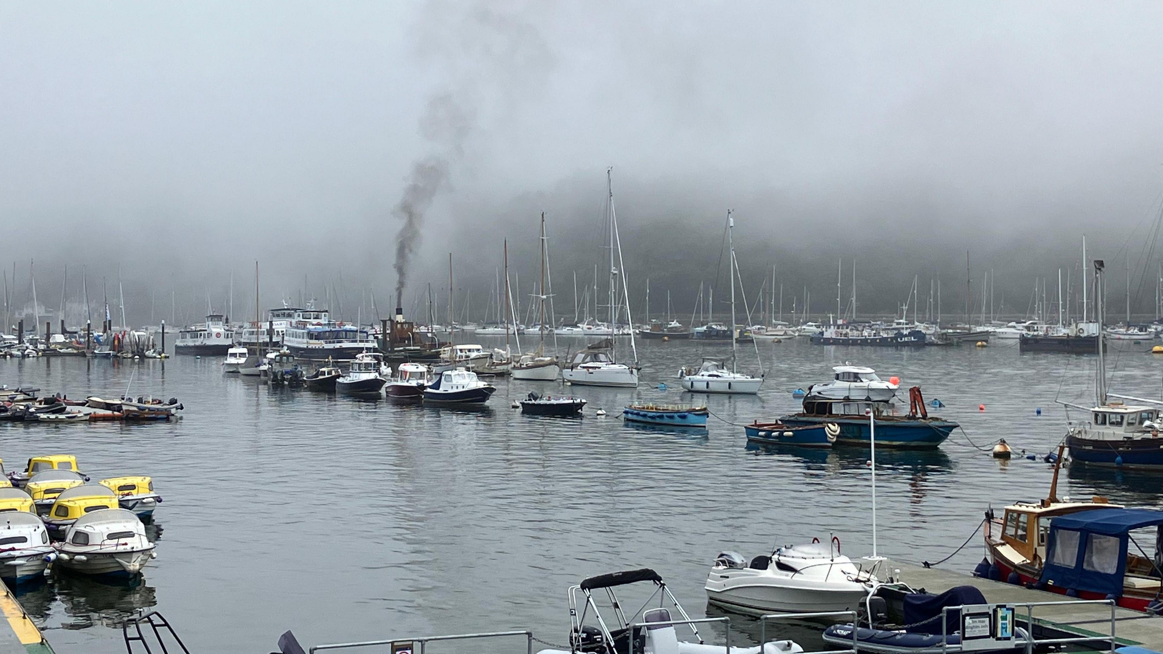 A harbour with rows of boats. There are some sailing boats, some are engine powered. The background is foggy. 