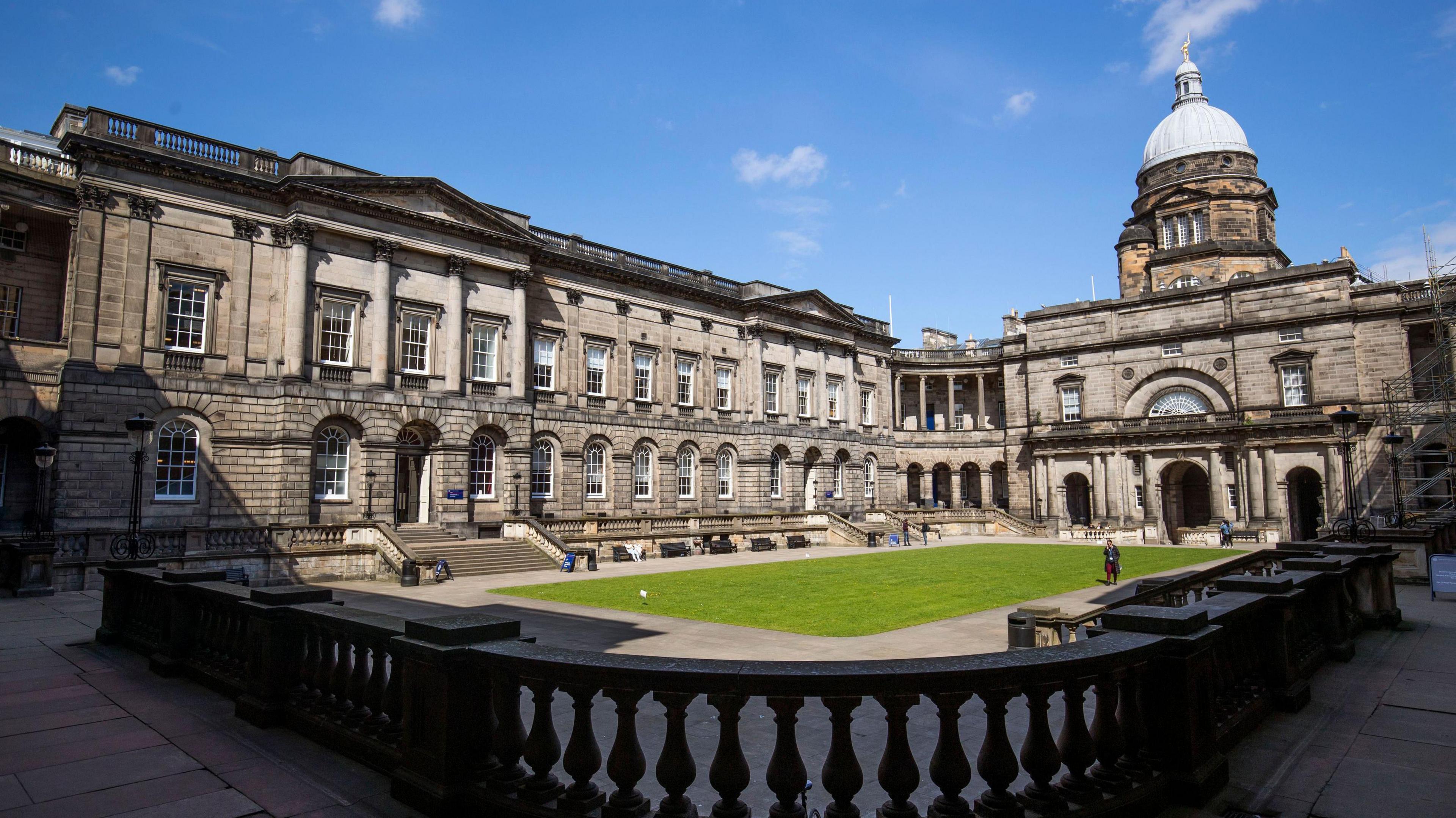 A general view of Old College at the University of Edinburgh. The sky is blue with a few scattered clouds. There is a rectangular patch of green grass in the middle of a courtyard. Older buildings surround the grass. On top of the one to the right of the image is a light-coloured dome.