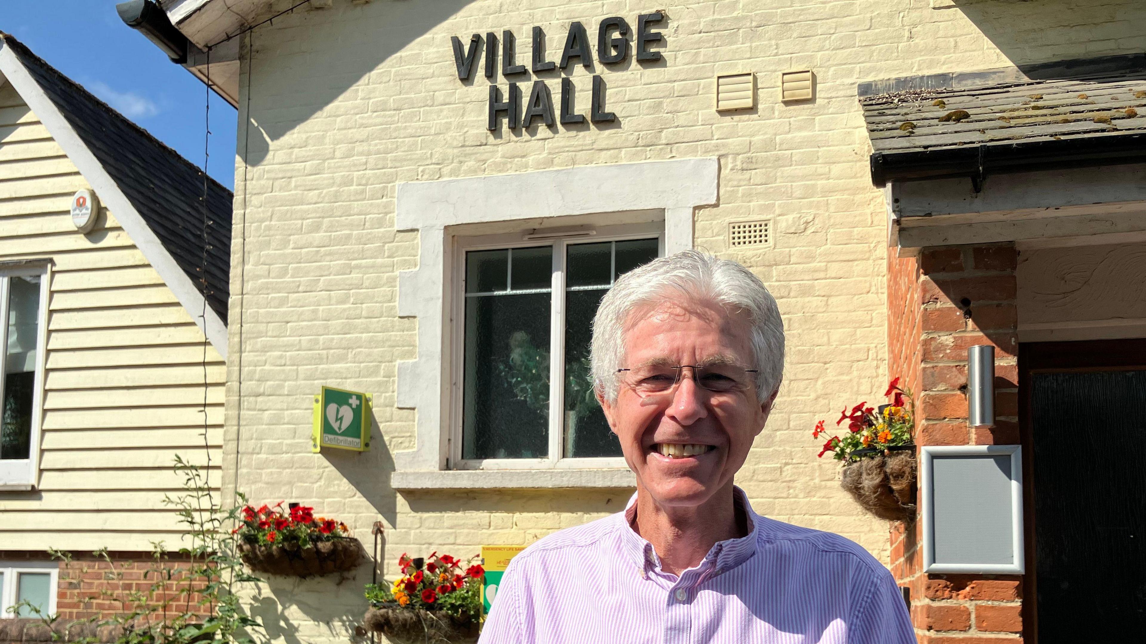 Alan McKenzie from Stop Wethersfield Airbase Prison, wearing a pink-purple shirt and glasses smiling at the camera, standing next to a building that says "village hall"