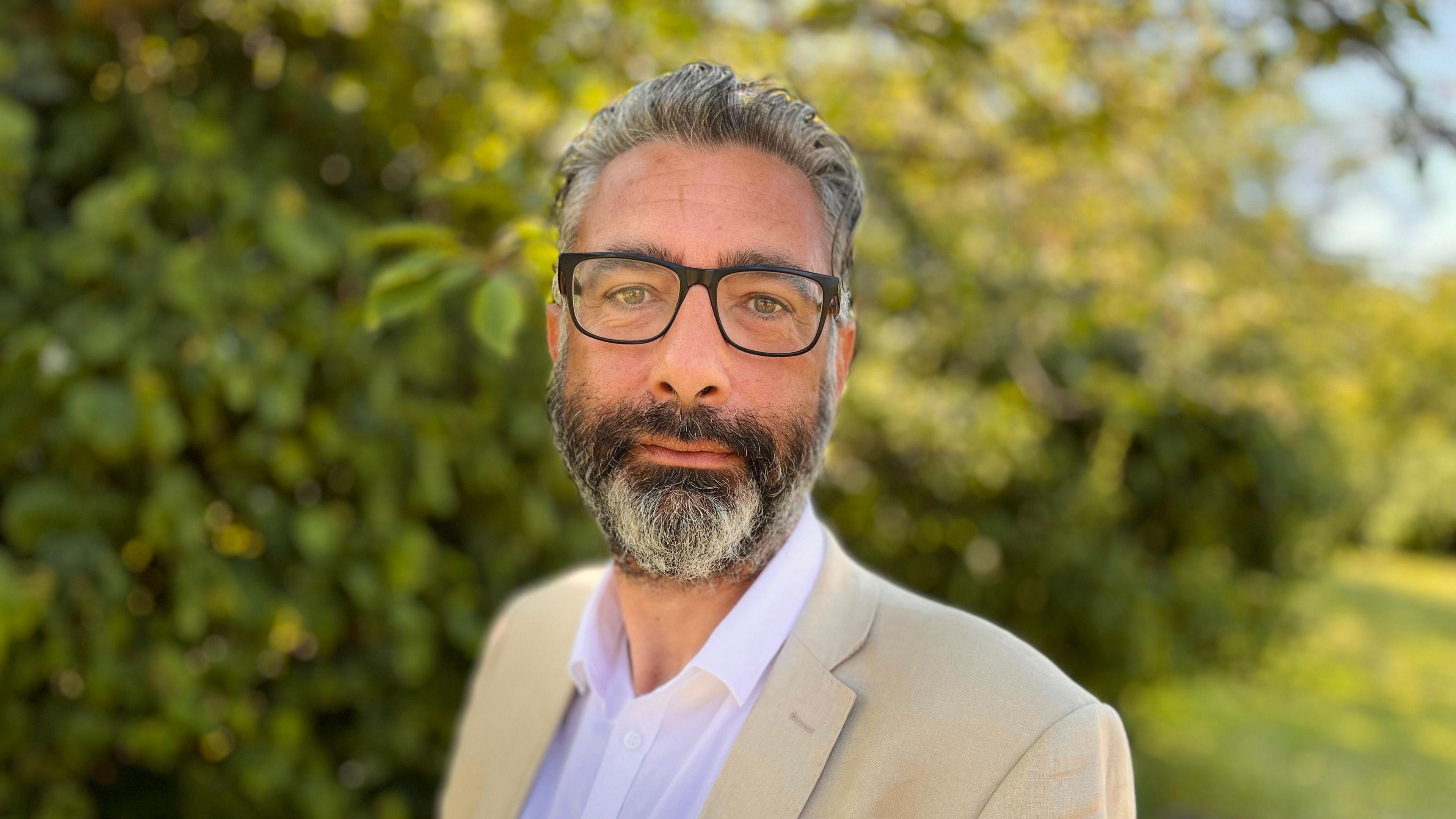 A head-and-shoulders picture of Asher Minns, who has grey hair and a beard and is wearing glasses, a beige jacket and an open-necked white shirt.