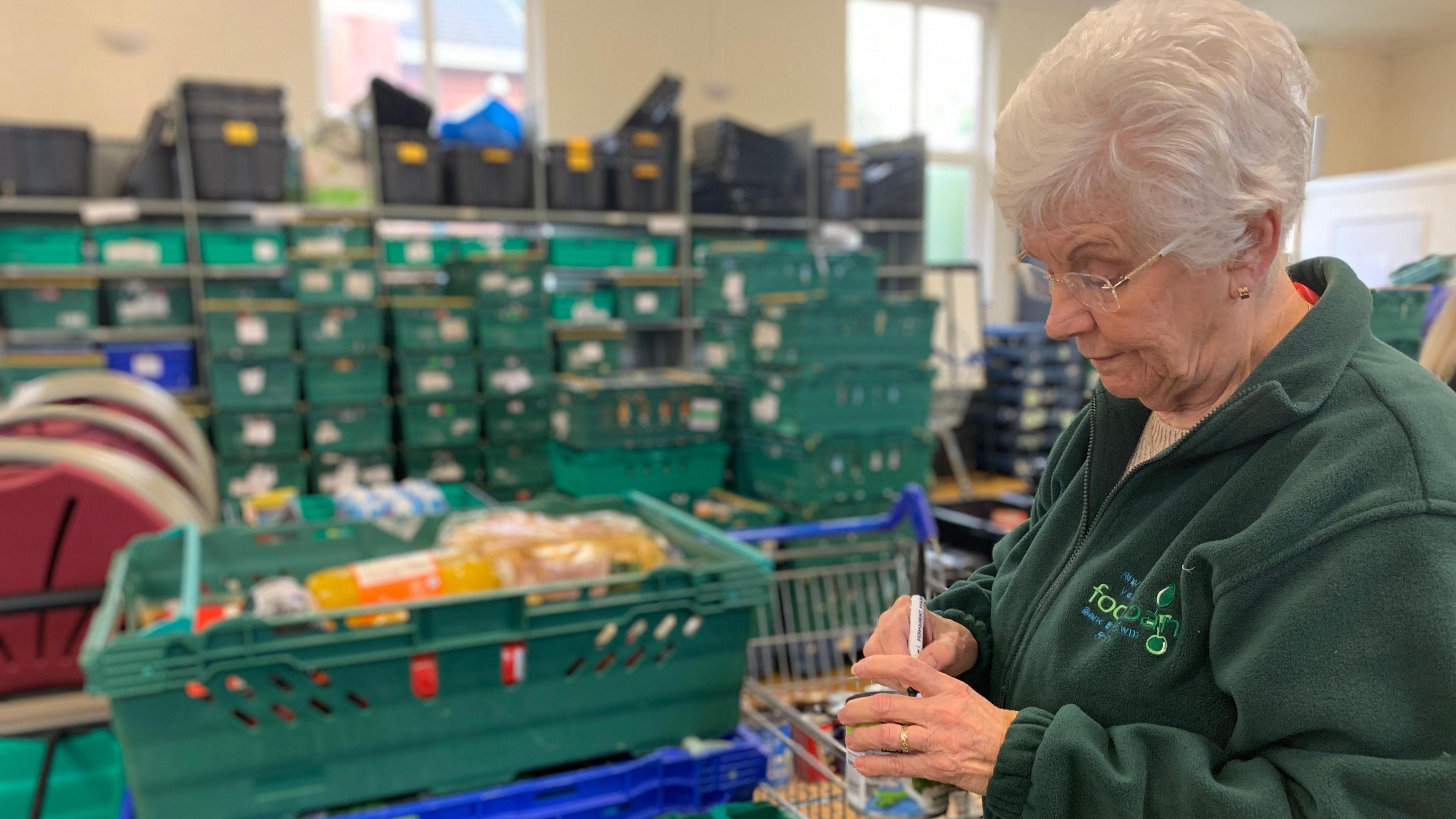 A woman is writing on a can. She is in a foodbank and there are crates of food to the left and behind her. She is wearing a green jumper which says "foodbank" on it. She has short white hair and glasses. 