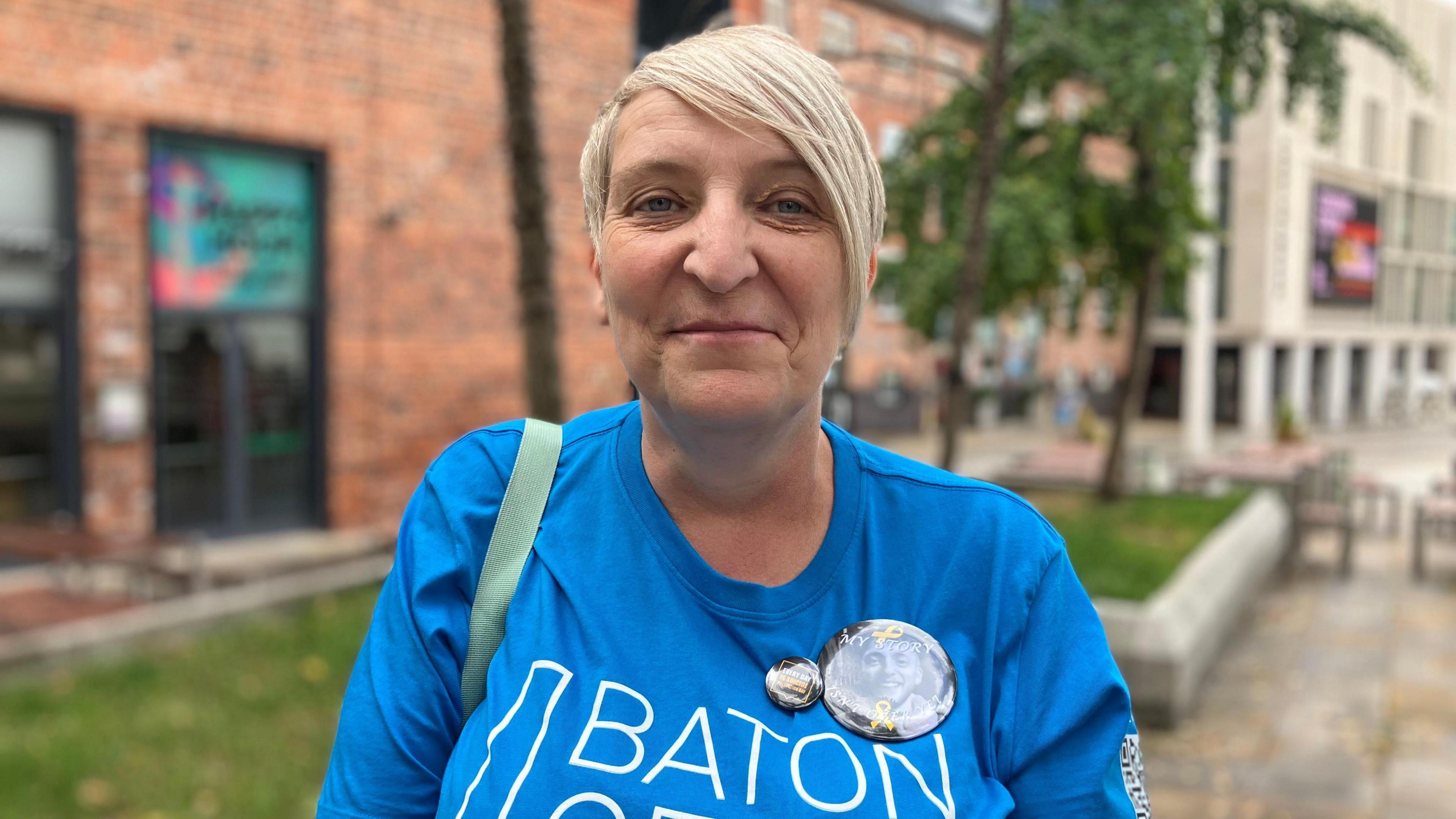 Trudie Stringer, who is wearing a blue t-shirt and badge with a photo of her son on. She has short blonde hair.