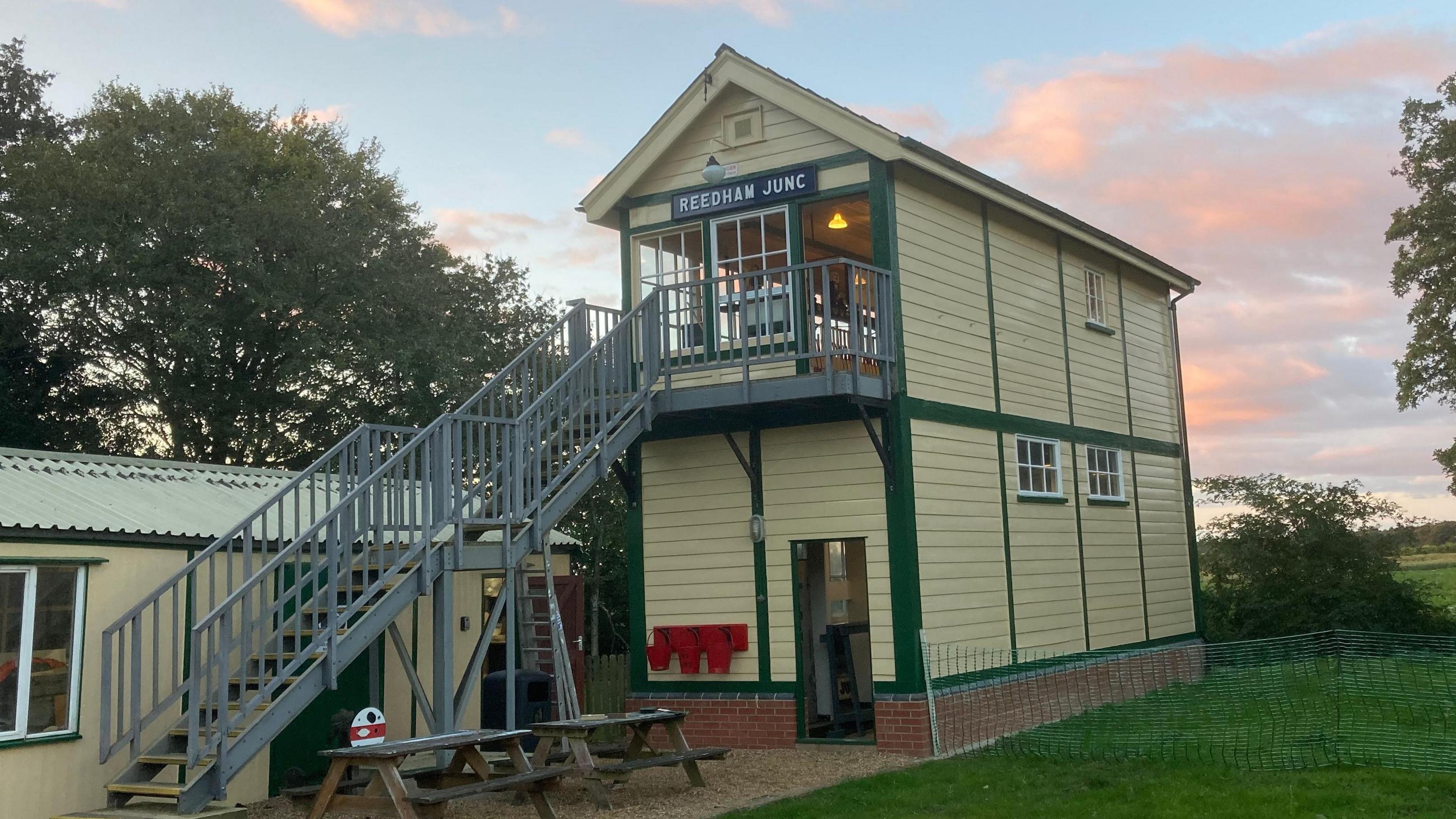 An external view of Reedham junction signalling box, a two-storey yellow building with grey metal stairs leading to the top floor