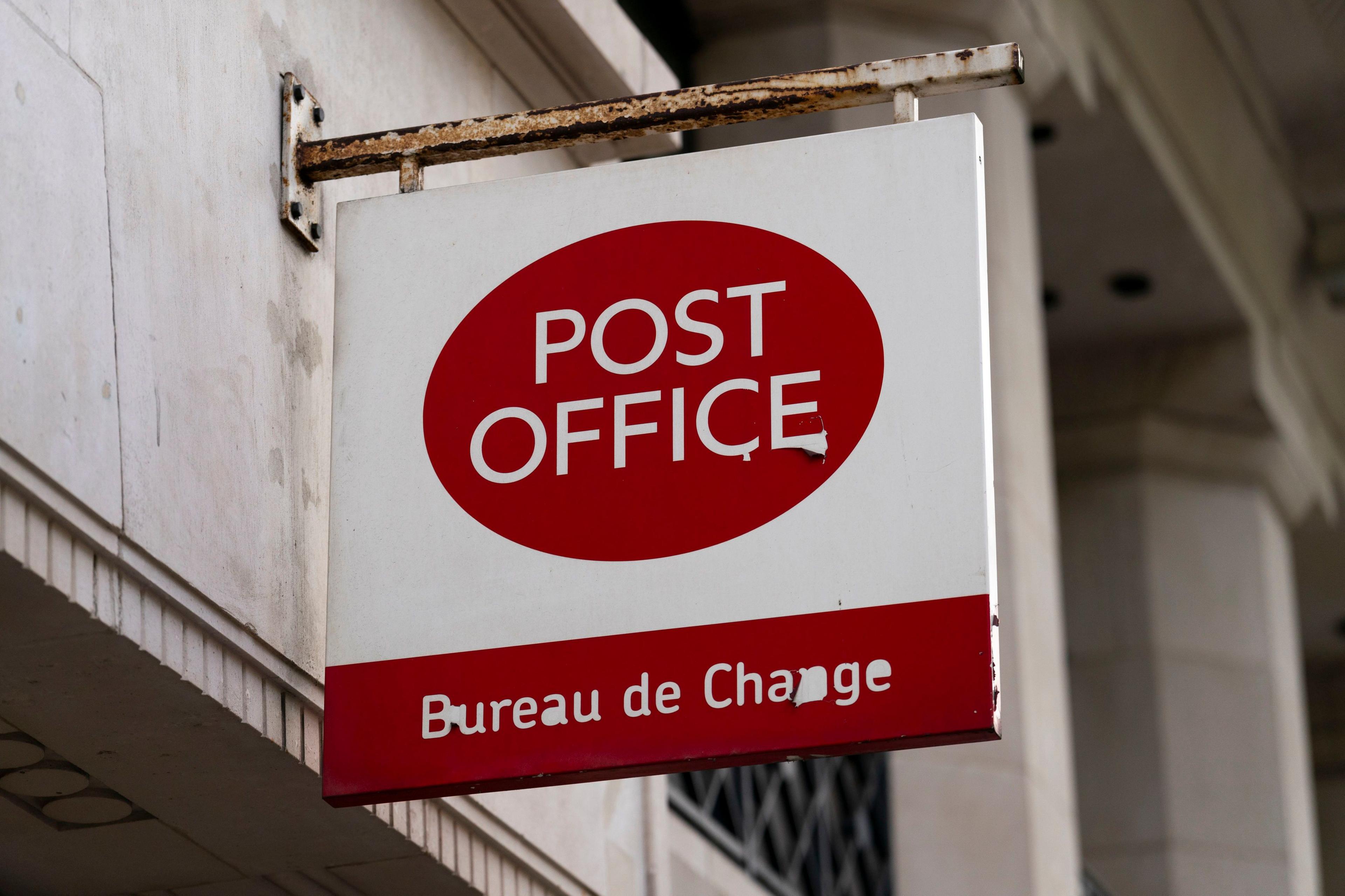 A post office sign on a building. Post Office is written in white writing on a red oval background and at the bottom of the sign is a red block with the words Bureau de Change in it