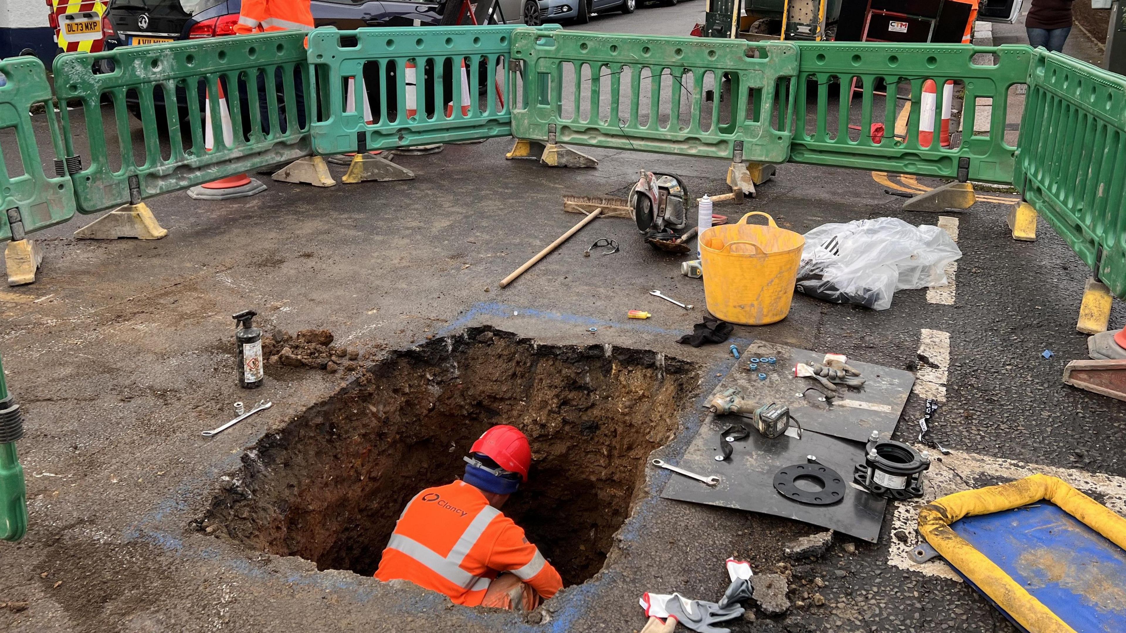 A man in orange hi-vis is working in a square hole dug into the road. The hole is cordoned off by green fencing. 