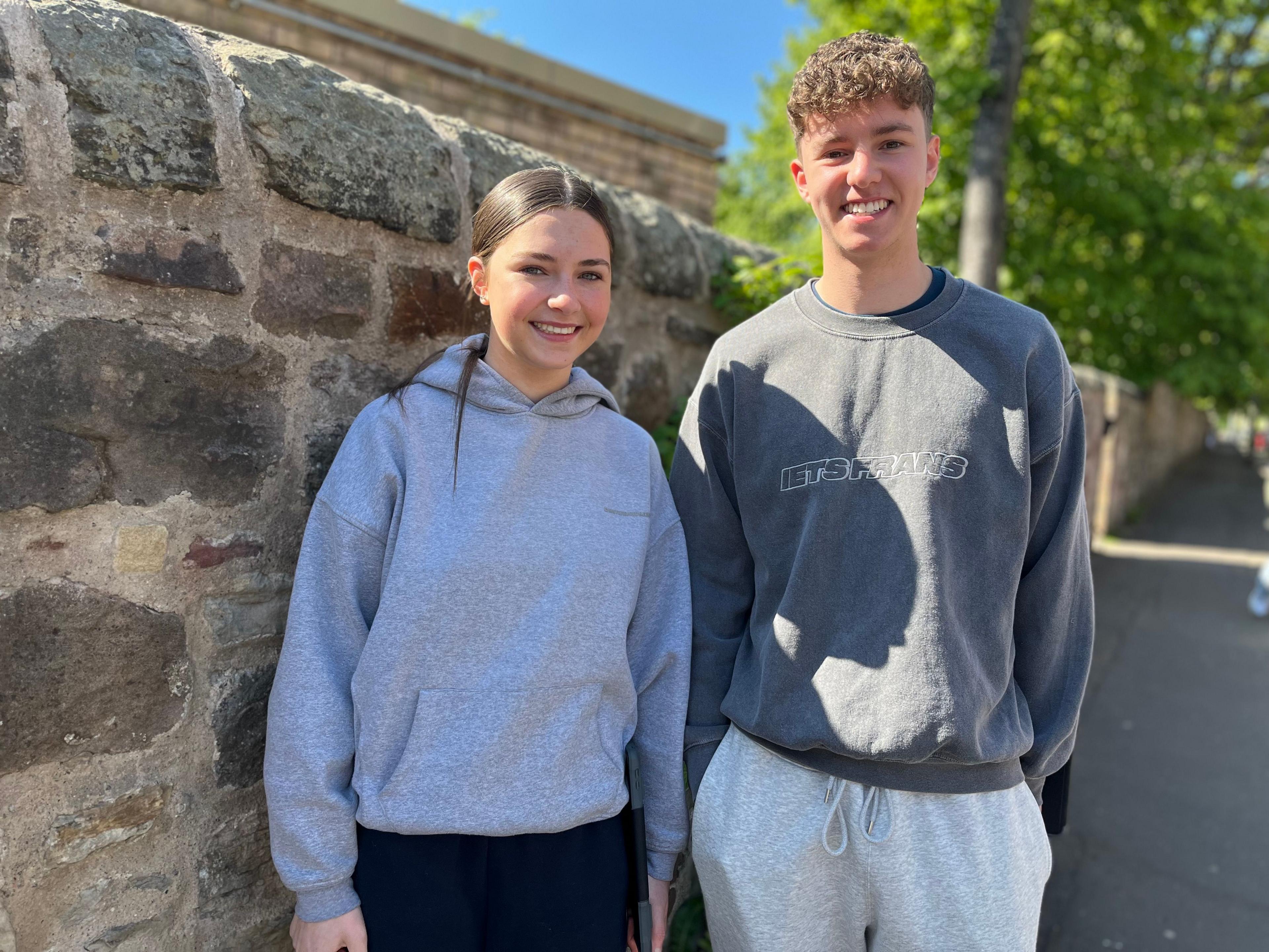 A girl in a grey hoody and a boy in a grey sweatshirt are standing by a grey wall, smiling.