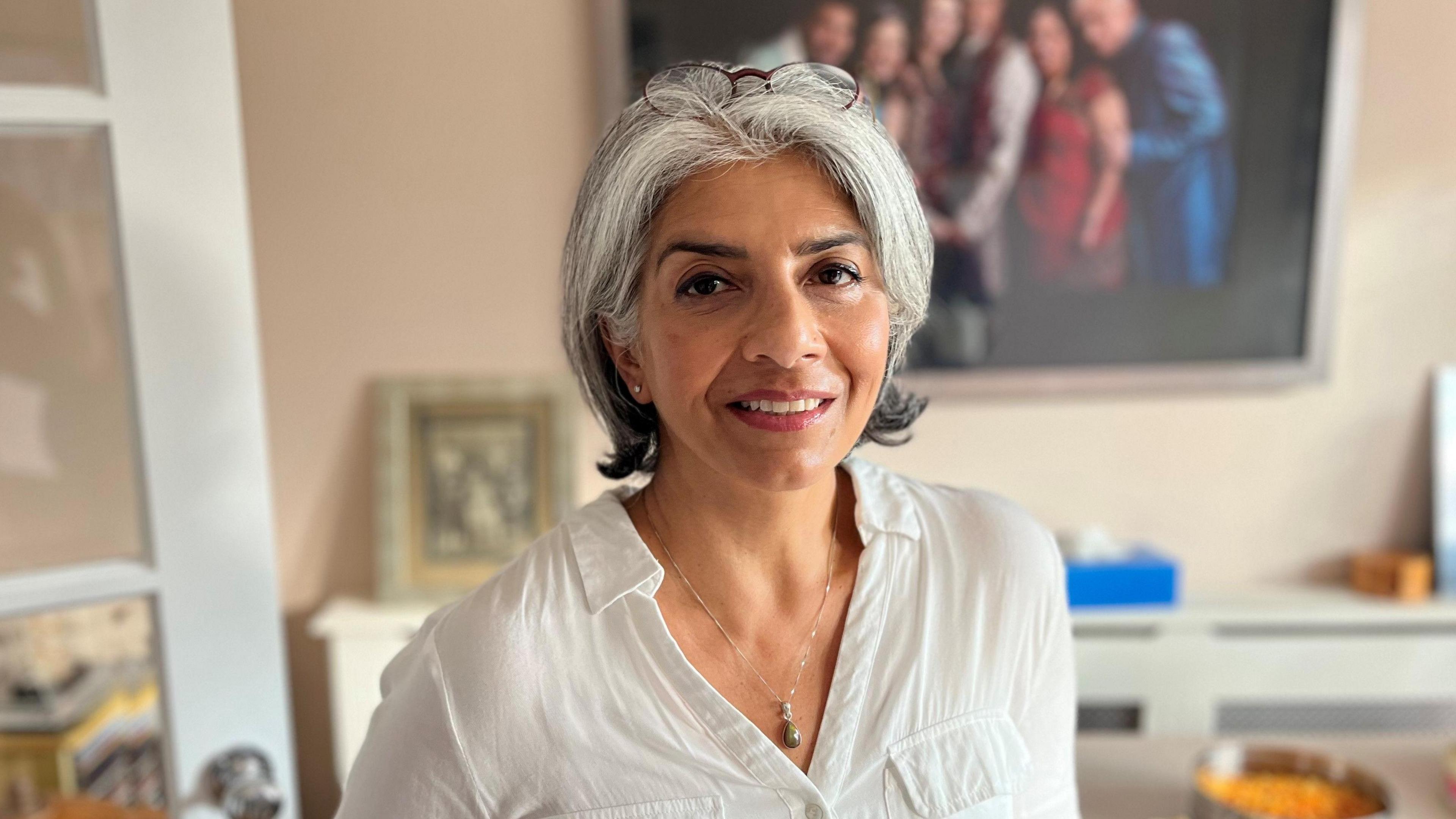 Pragna Hay wearing a white top, with shoulder-length grey hair. She has reading glasses perched on her head. She is wearing a necklace with a pearl pendant on it. She is standing in her lounge and behind her are a couple of family photos.