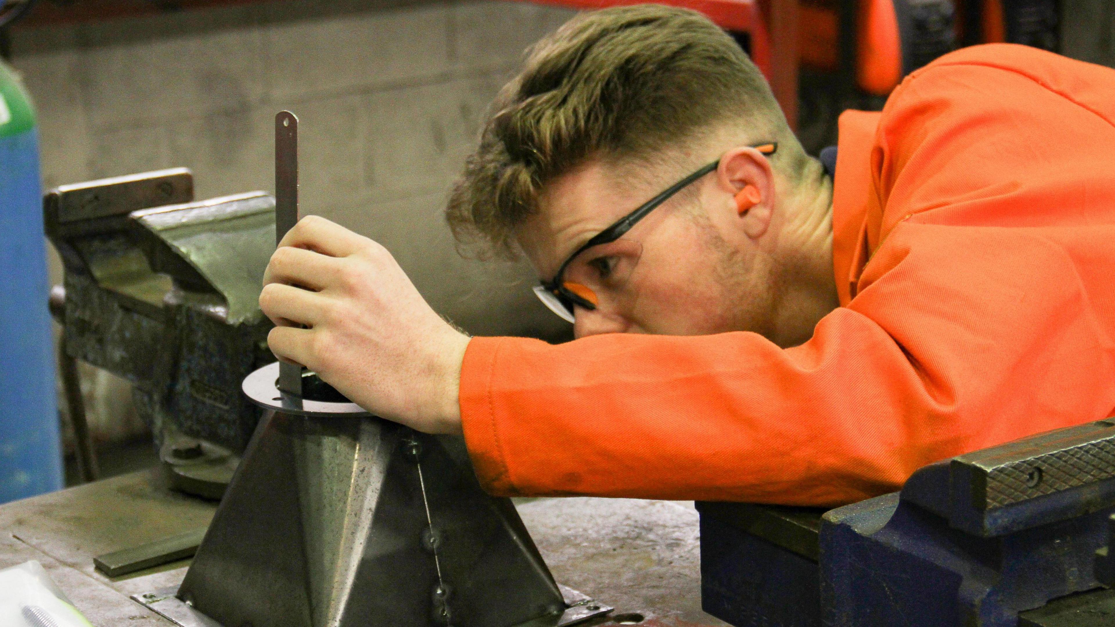 A student wearing a bright orange jacket and protective goggles works on a piece of machinery.