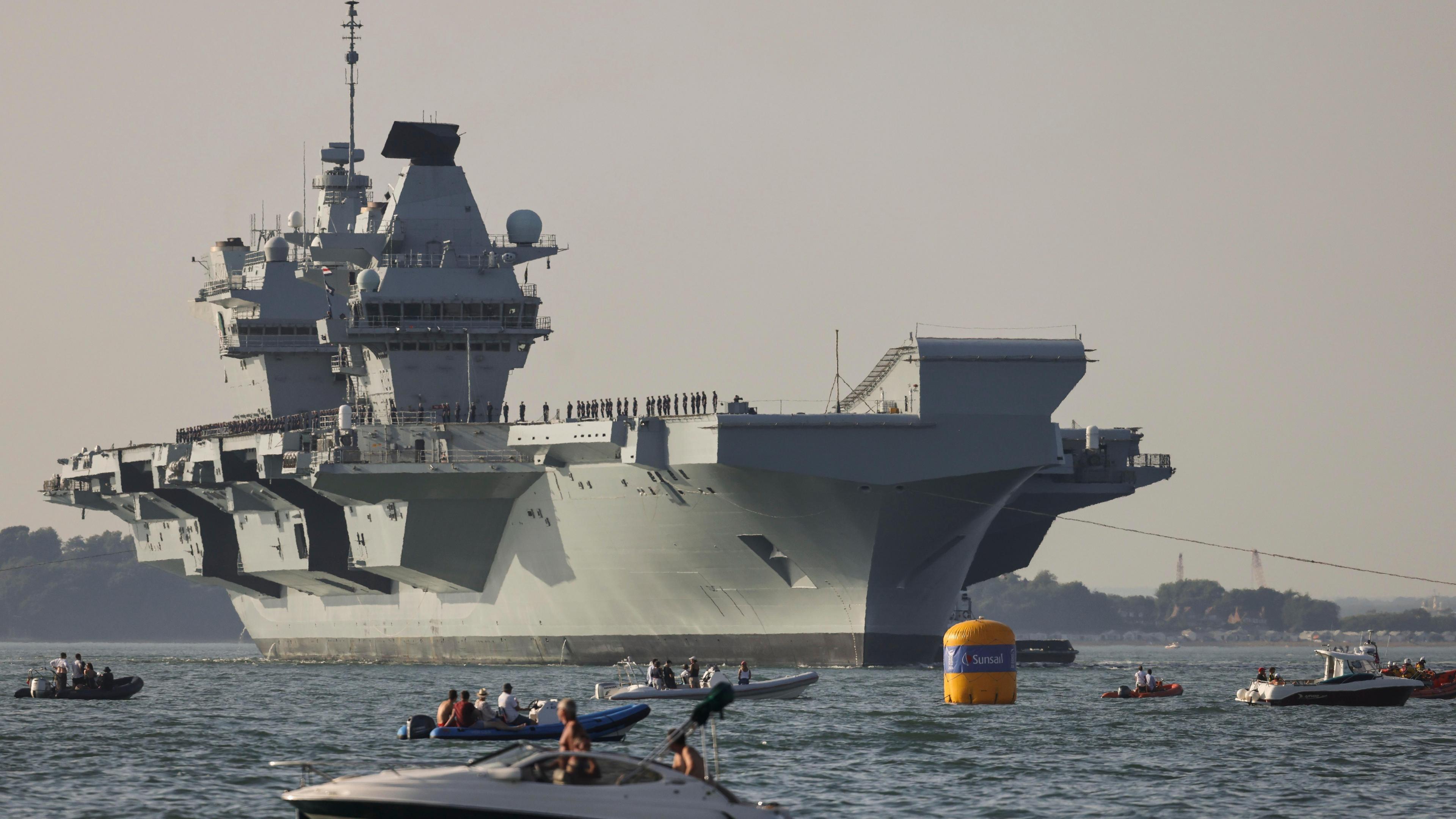 Queen elizabeth on the water, with lots of smaller boats in front of it. The sky is orange.