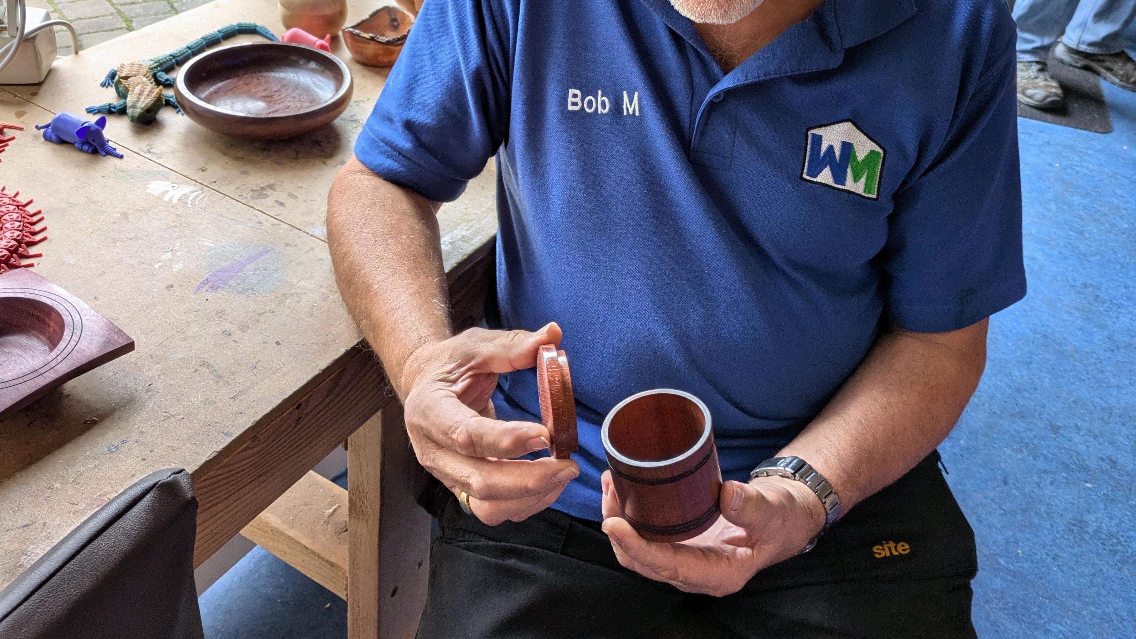 A man pictured from the torso down opens a small wooden beaker, holding the lid in one hand and the beaker in the other. He is wearing a blue polo shirt emblazoned with the name 'Bob M'.