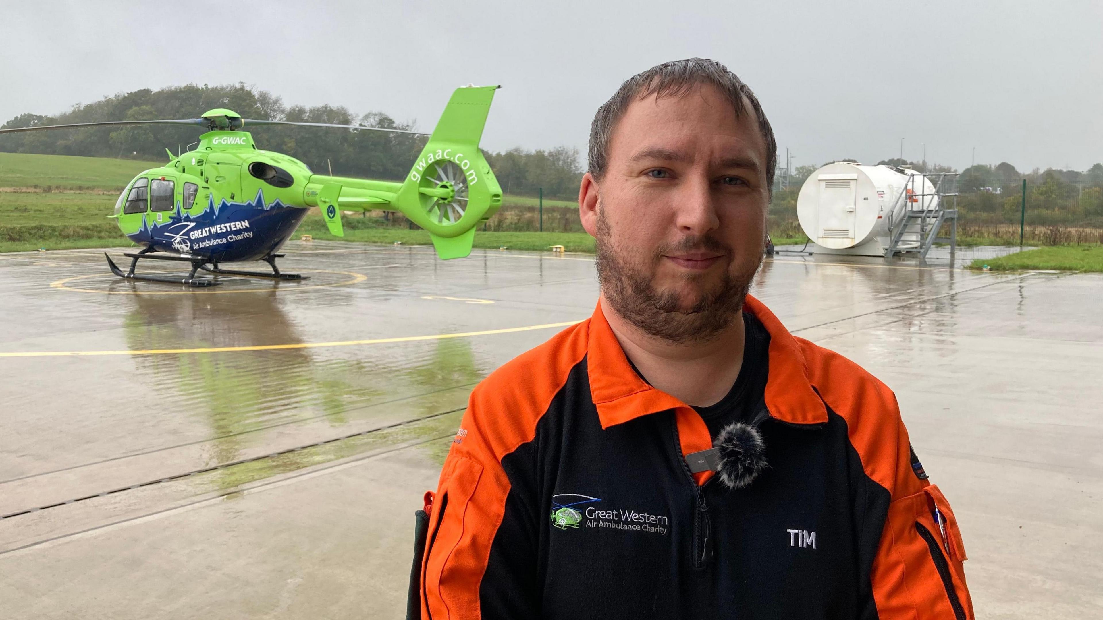 Tim Ross-Smith looking at the camera, half-smiling, in an orange and navy jacket, stood with a helicopter behind him on a helipad