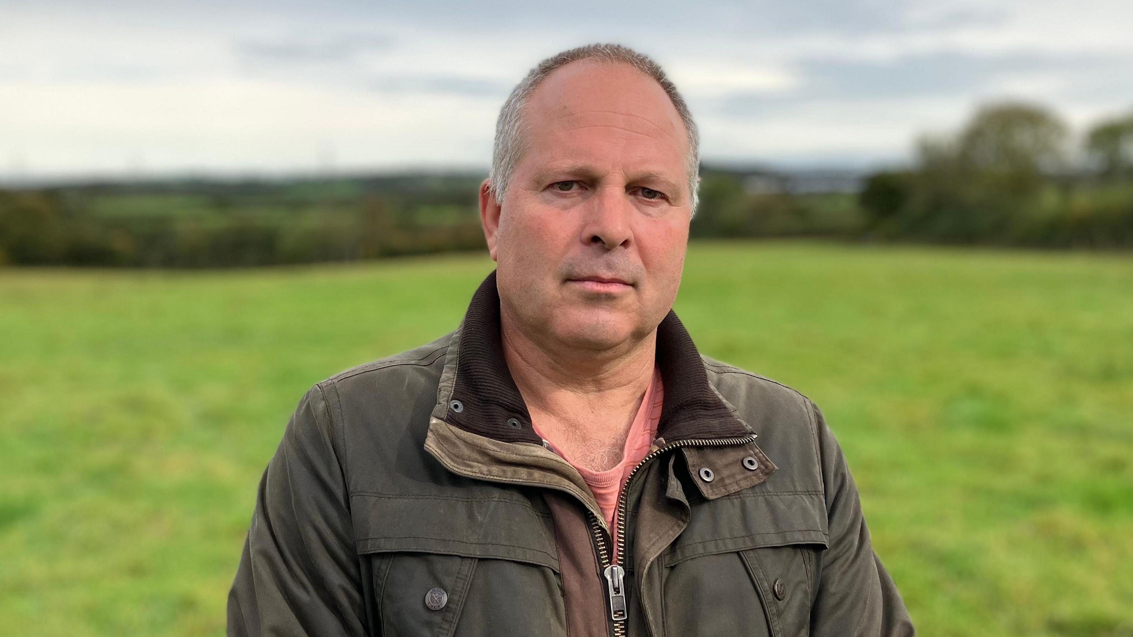 A man stands in a field in the countryside, there are trees and hedgerows in the background.