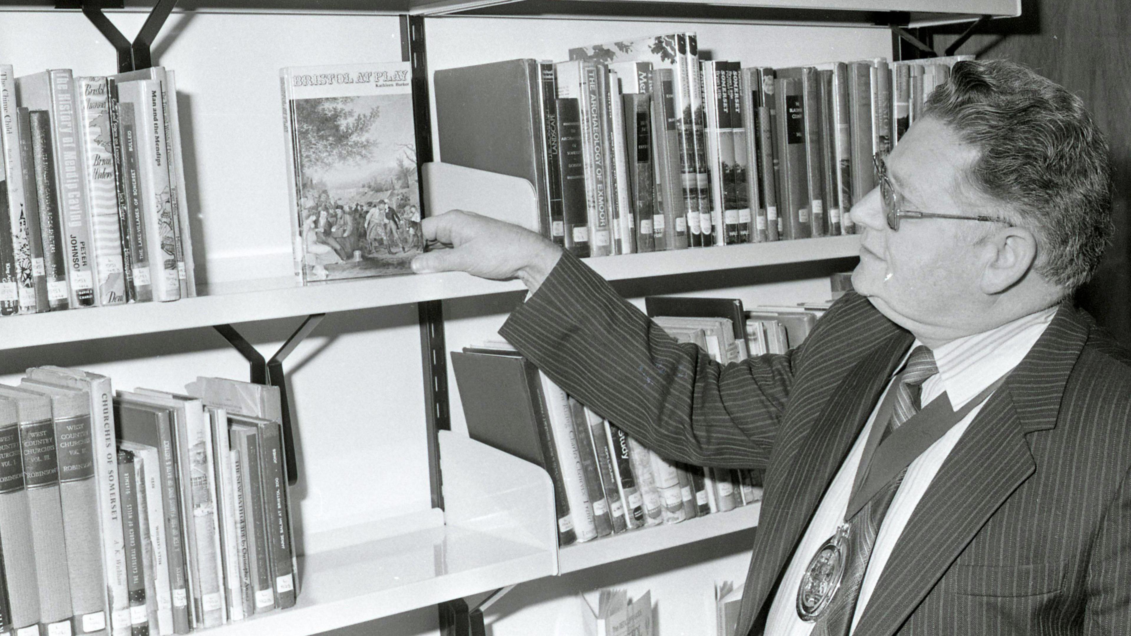 A black and white picture of a man wearing a pinstripe suit and glasses. He is looking at a book on a shelf admiringly, while other books are stacked beside it on the shelves using a dewey decimal system. 