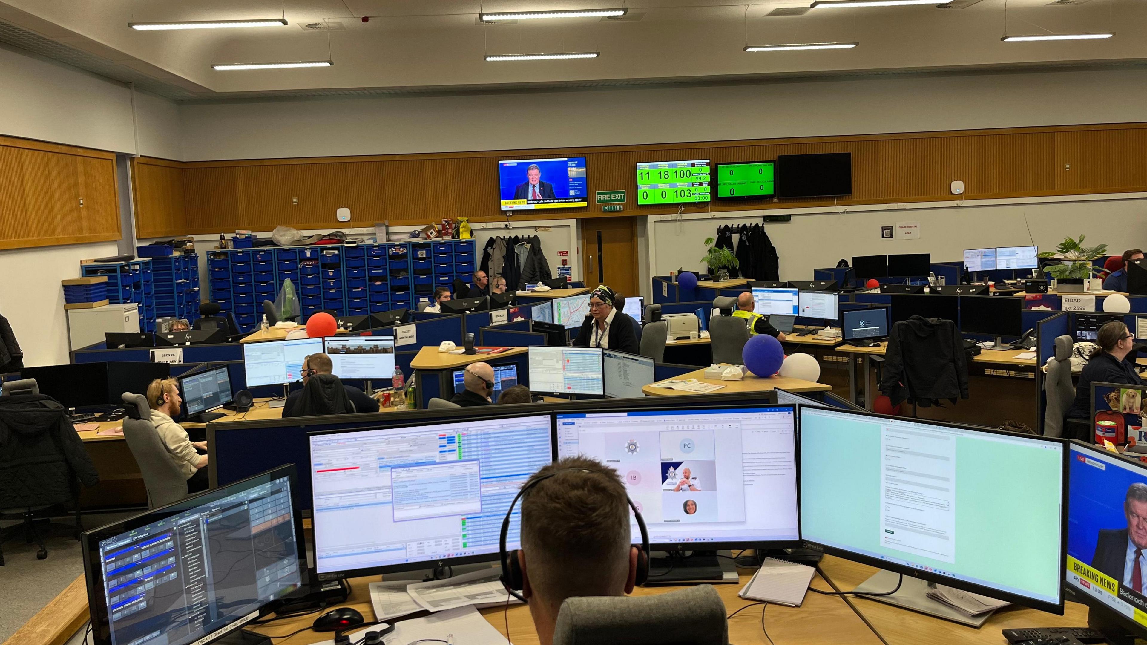 Part of a police call handling office shows many people sitting at desks, each of which have several monitors on them. There are lots of blue lockers at the back, against a wall, and coat hooks. High up on a wall is a large TV screen on a news channel and other monitors