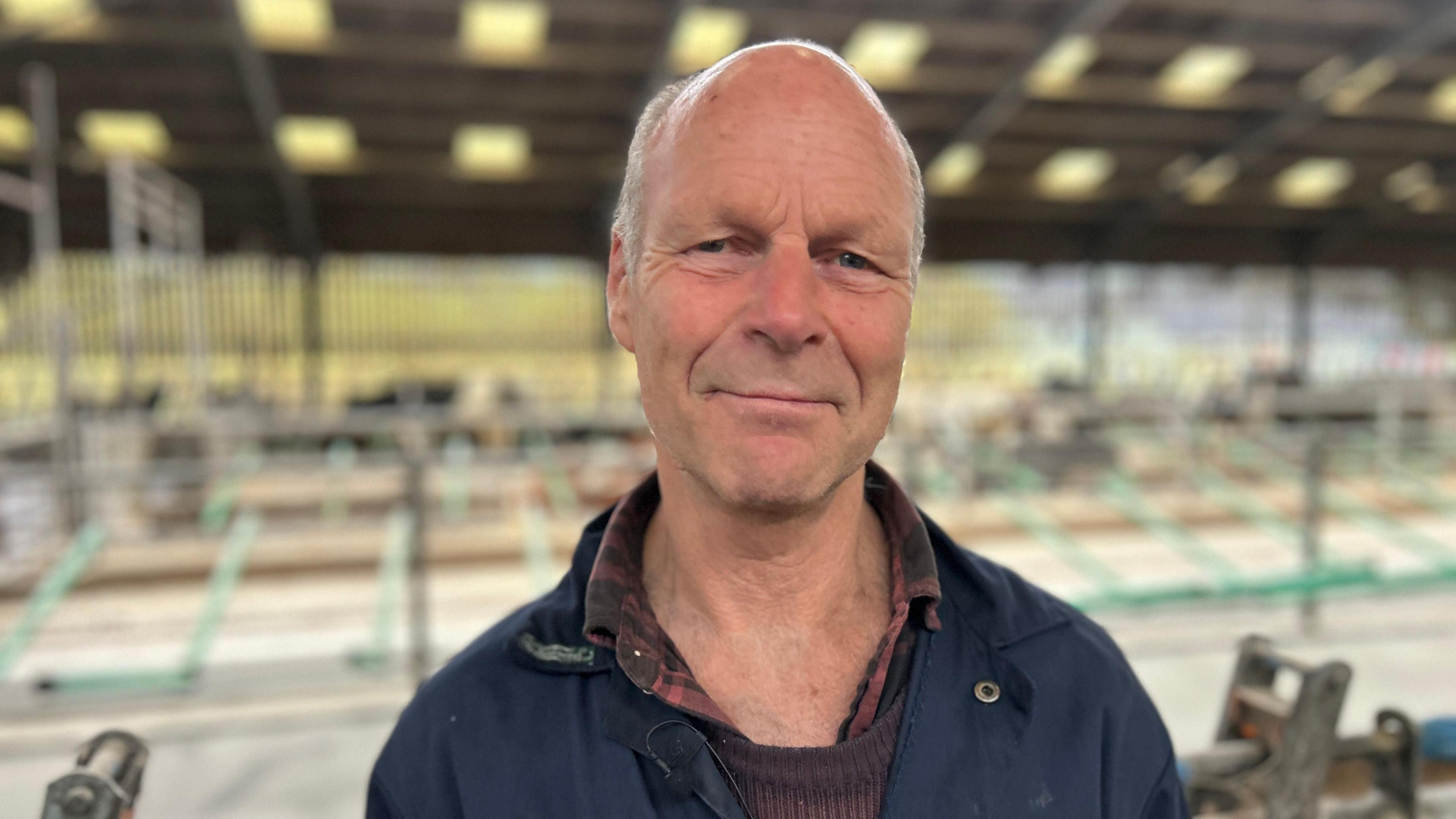 David Finlay wearing a blue boiler suit and burgundy shirt and jumper, smiling inside the cow shed