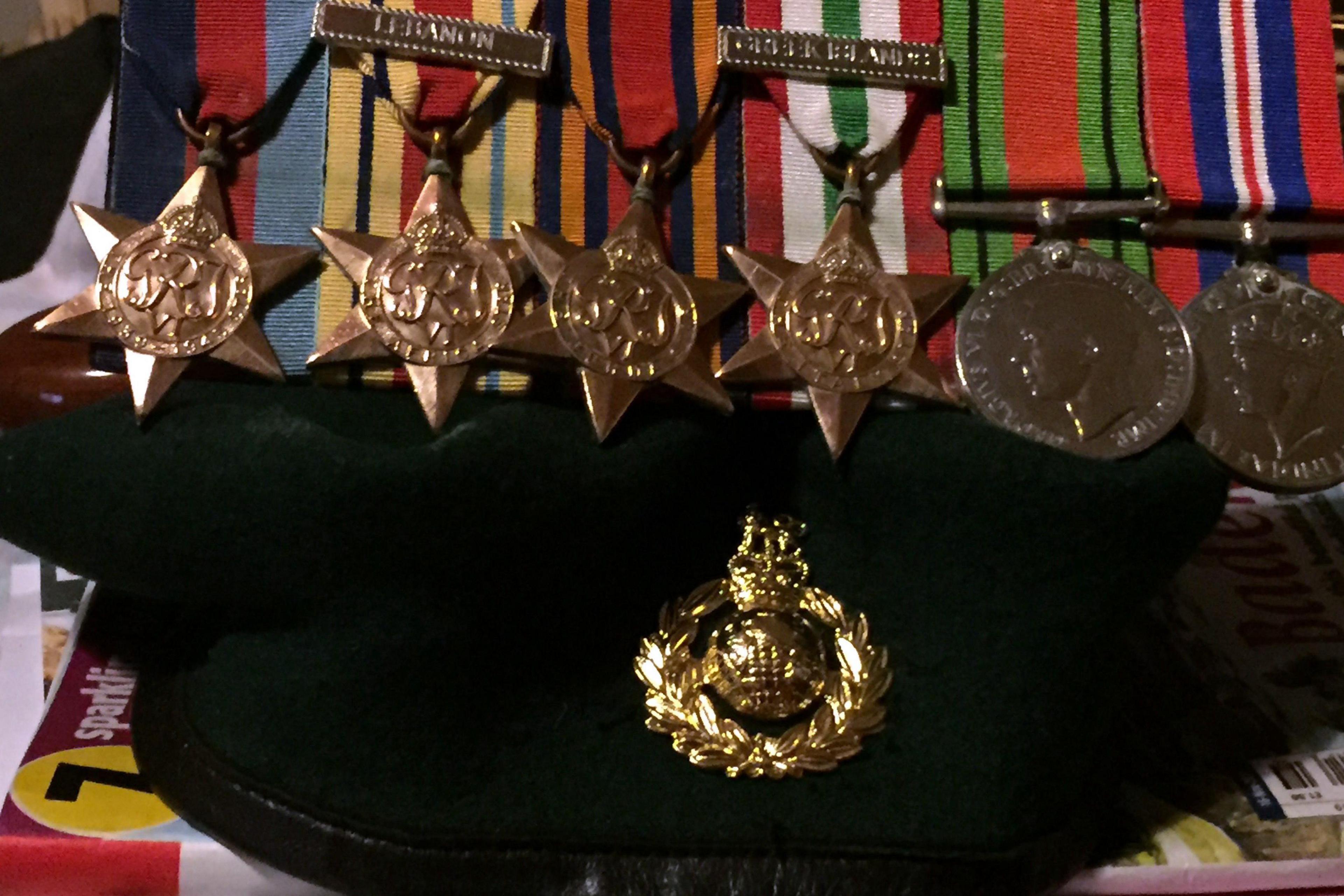 A close-up image showing six military medals laid out in a row, each attached to brightly coloured ribbons. Beneath them is a dark military cap bearing a gold coloured insignia