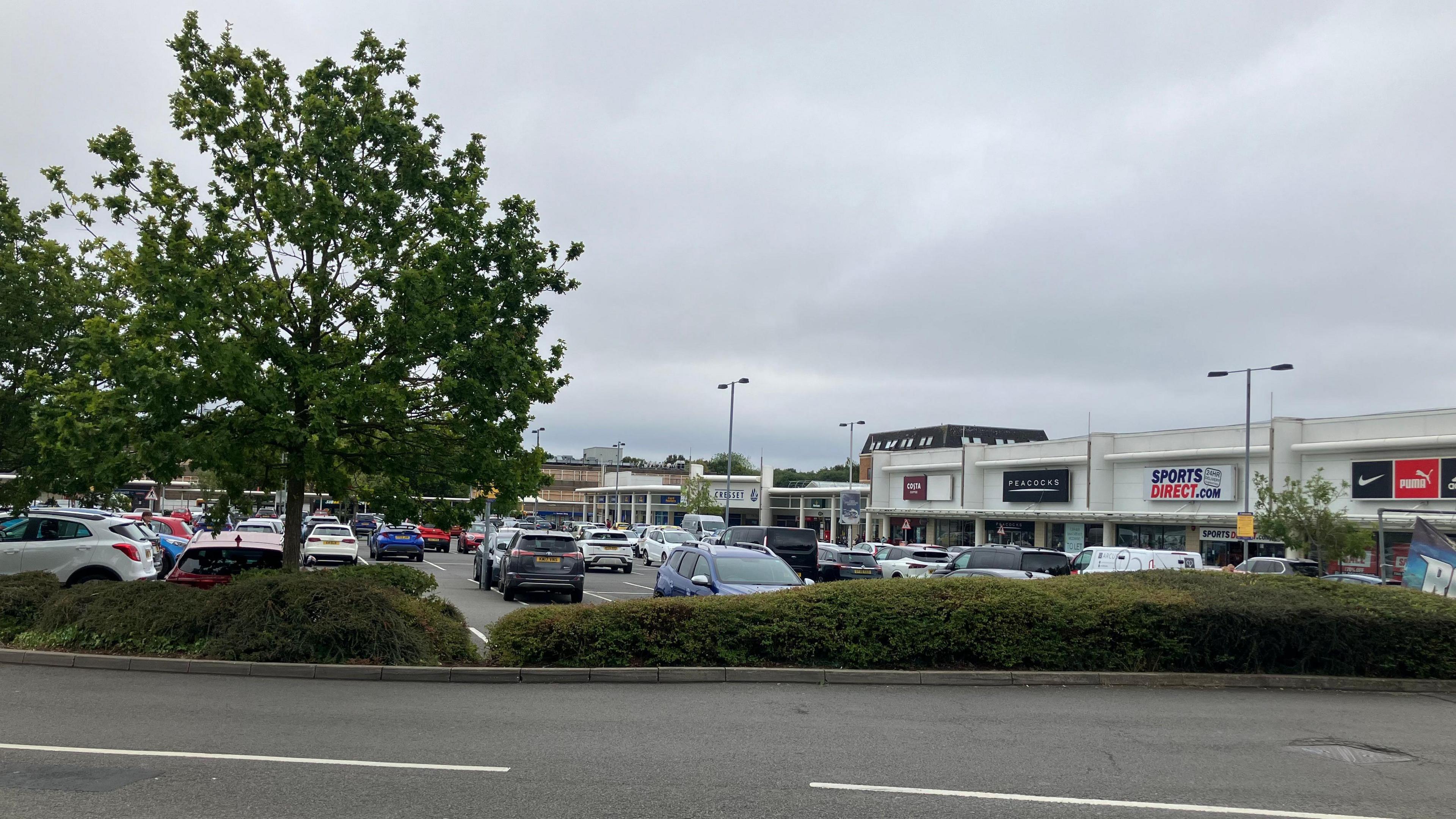 In the foreground, a stretch of road, separated by shrubs and a tree from a car park filled with parked cars. On the right is a line of shops, including Sports Direct, Peacocks and Costa Coffee