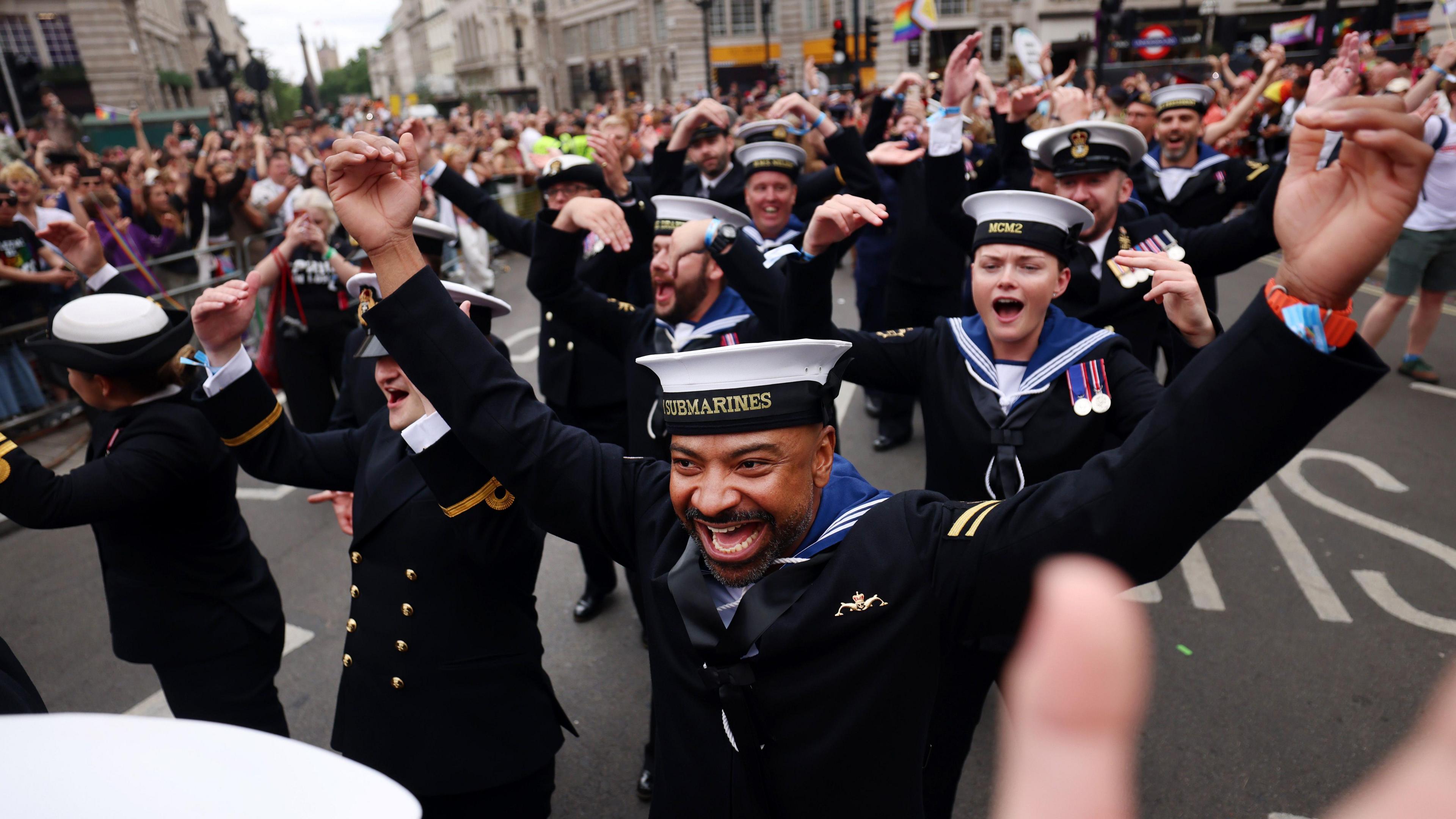 Royal Navy sailors in uniform march in the parade - several of them have their hands in the air and are cheering. 