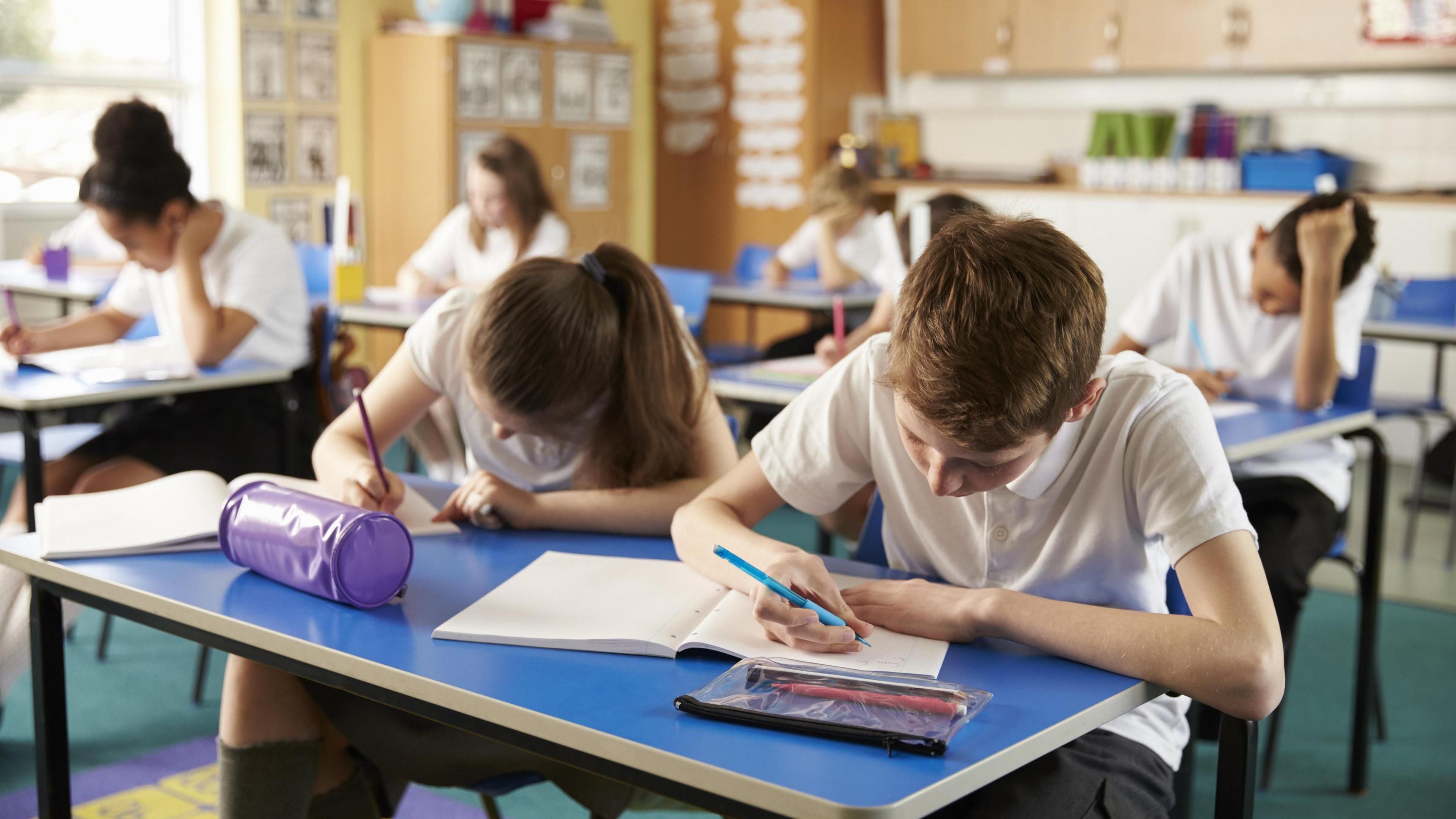 Children sitting at blue desks in a classroom. They are wearing a school uniform of white collared shirts and dark trousers or skirts. There are papers and pencil cases on the table, and the children have their heads down, focusing on their work.