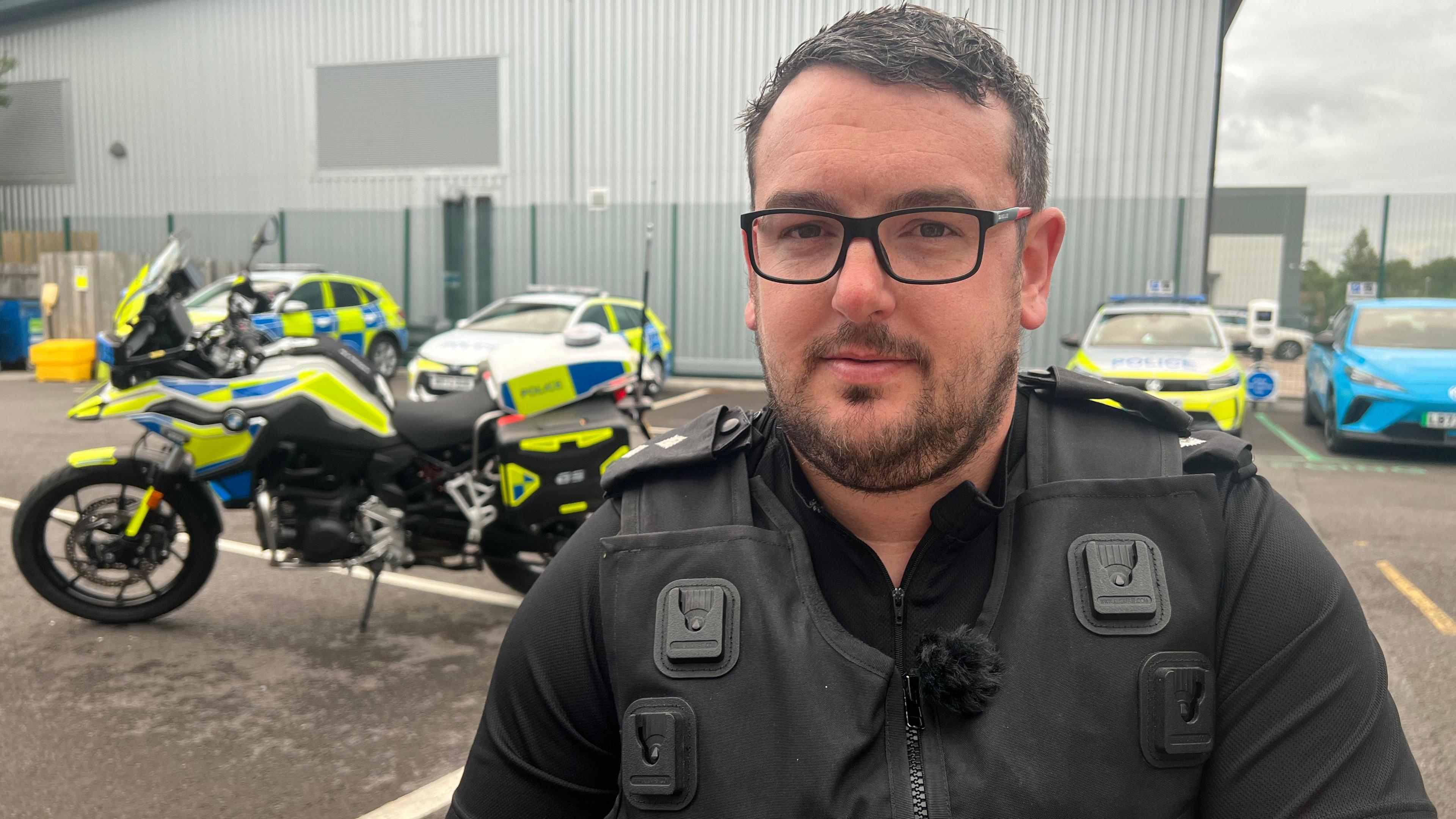 Matt wearing his police uniform and black rimmed glasses, with short dark hair smiling at the camera. He is standing in a police car park with police cars and a police motorbike in the background.