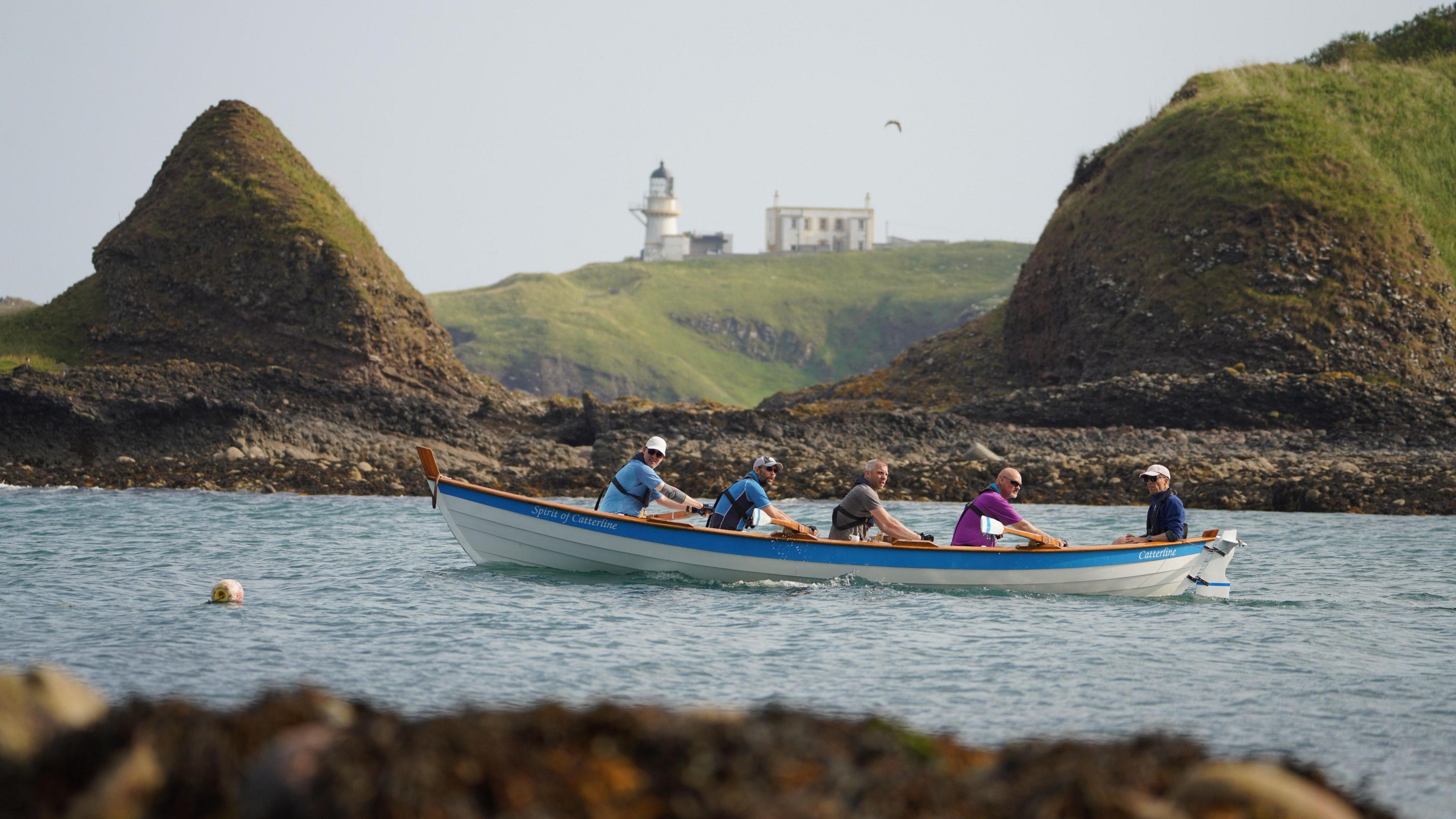 A group of five men, wearing brightly coloured T-shirts in a rowing boat going past some green rocks. In the distance on a hill is a lighthouse and another white building.