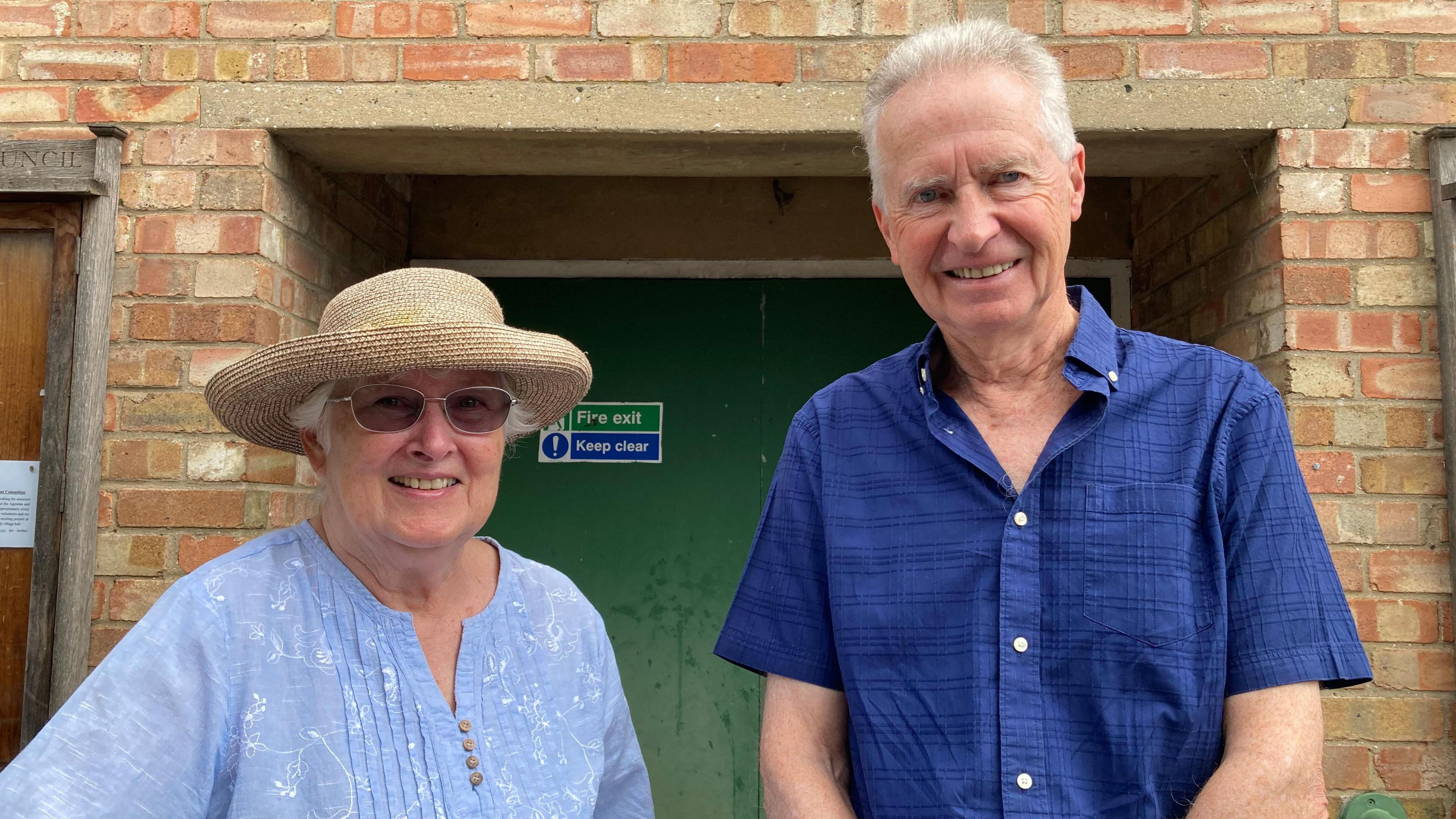 Ruth White and John Fisher are smiling at the camera and standing in front of a door leading to a village hall. She is wearing a light blue blouse, a straw sunhat and sunglasses. He is wearing a dark blue shirt and has short, grey hair.