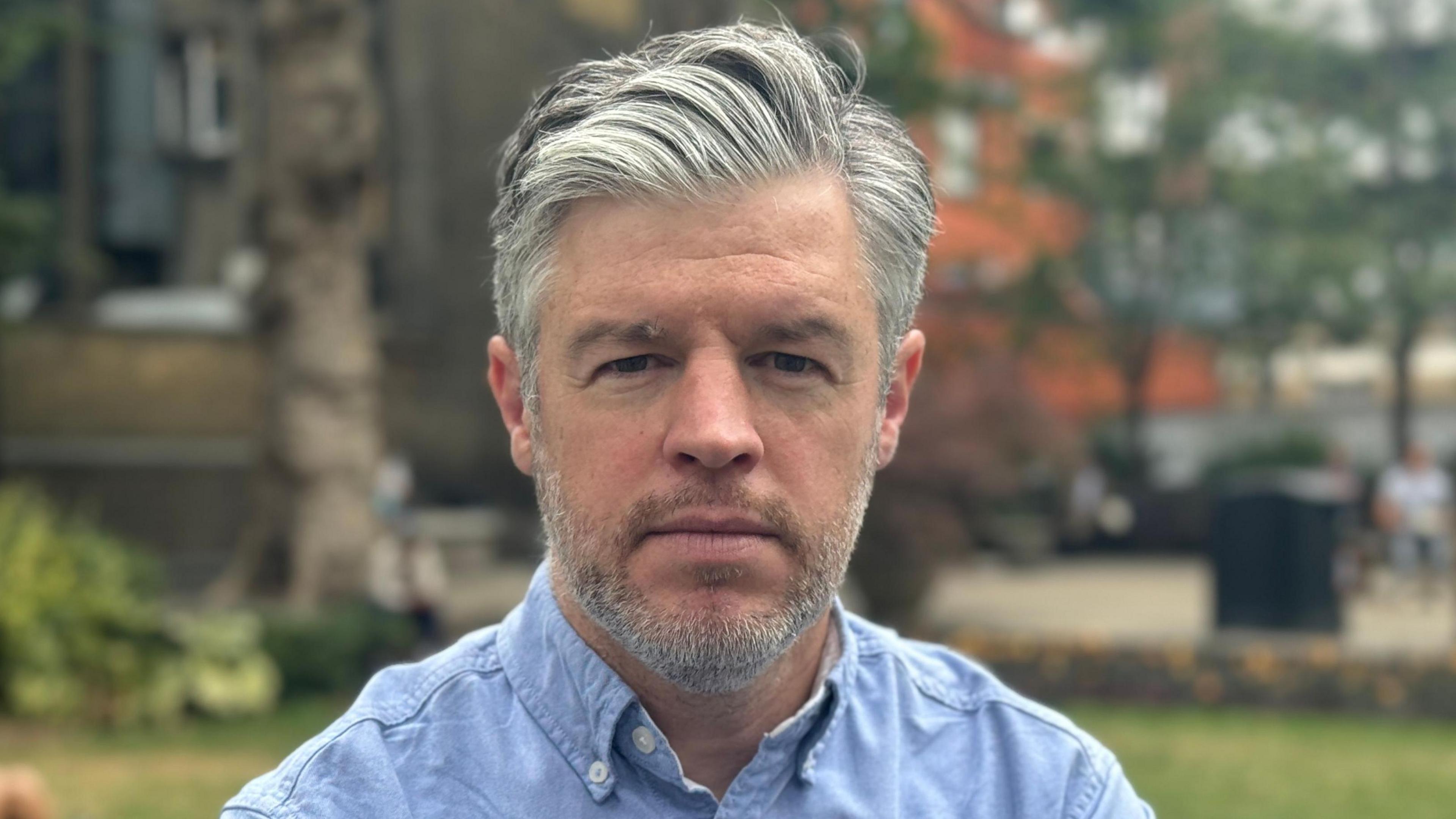 A man with grey hair worn in a side-part looks at the camera. He is stood in a park and is wearing a blue shirt with the collar unbuttoned.