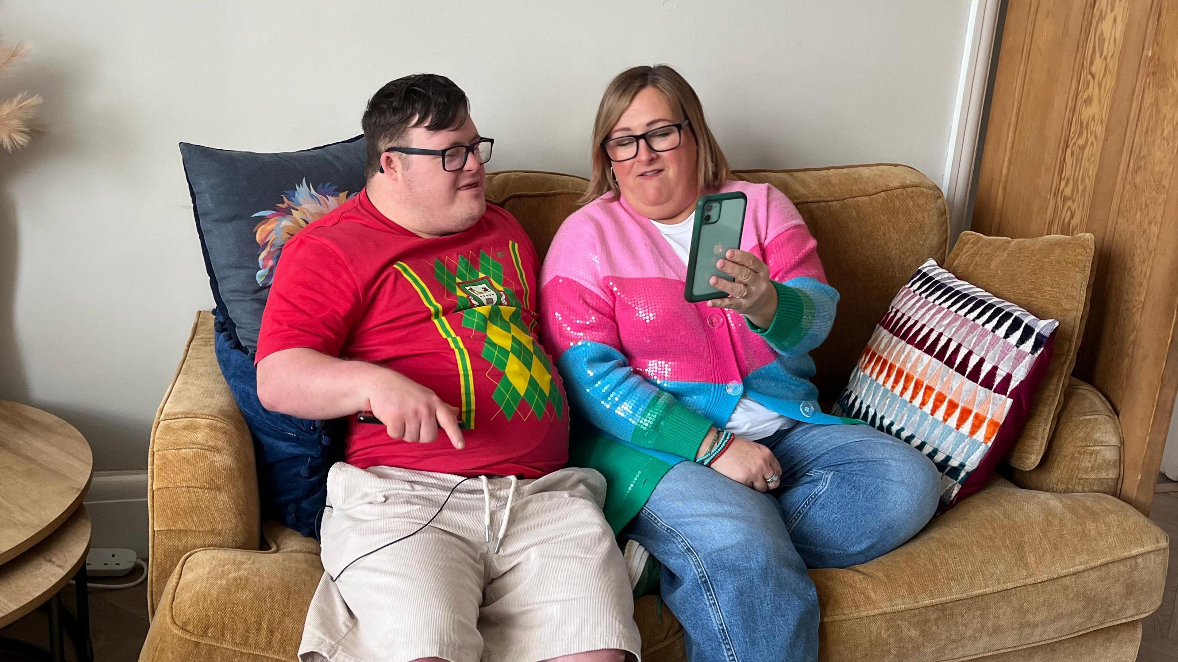 Joel and his mum Clare sitting on a grey sofa, facing each other and laughing. Joel has Down Syndrome, has short brown hair and is wearing a red, green and yellow t-shift and dark-rimmed glasses. Clare is wearing  a pink, blue and green sequined cardigan and has a blonde bob.  