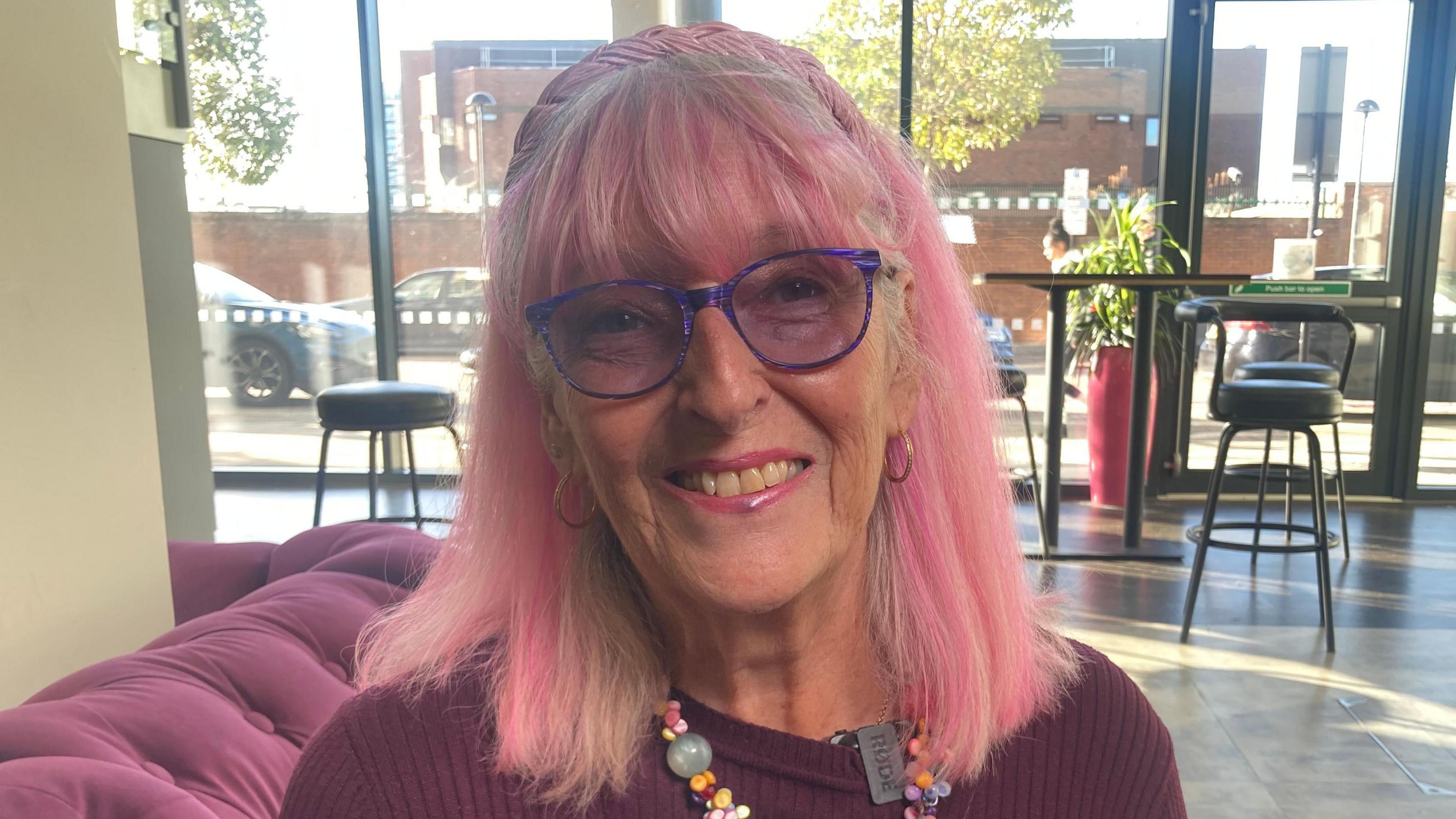 Lynn Fox is smiling at the camera, she has white and dyed pink hair, is wearing a red jumper, and is sitting in a cafe environment.