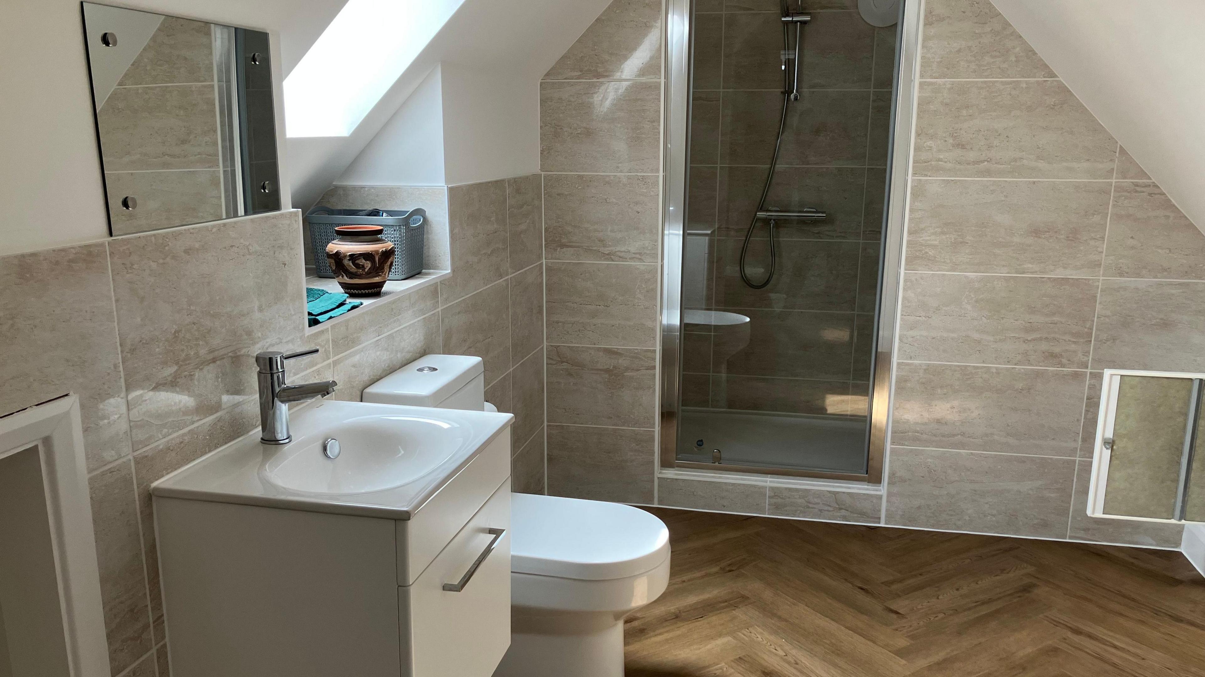 A clean en ensuite bathroom with grey tiled walls, a white sink and toilet, mirror and shower built into the wall. The floor is wood panelled. There is also a sky light above the toilet and a small vase on a windowsill underneath it. 
