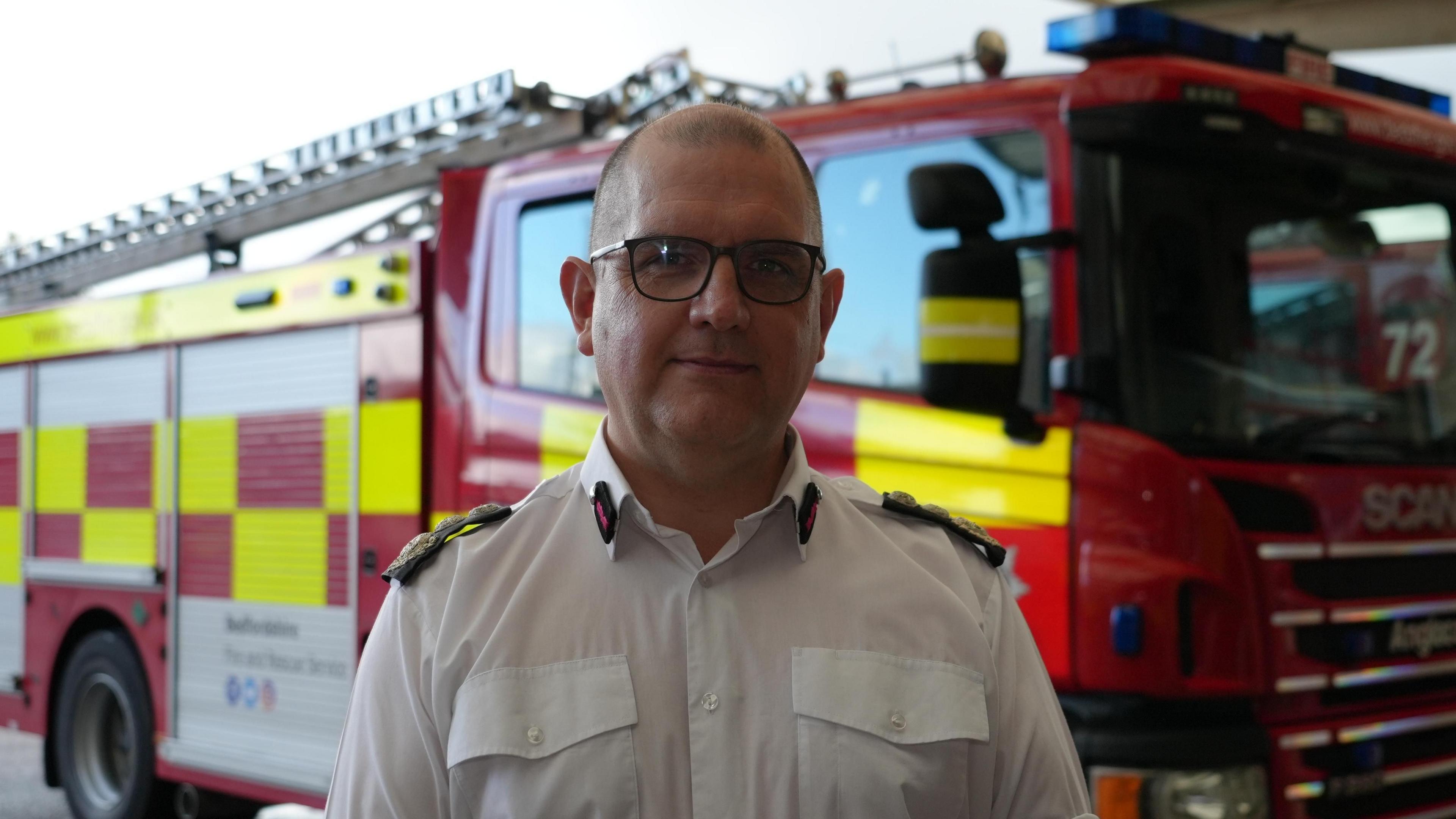 A balding man wearing glasses with a white fire service shirt standing in front of a fire engine.
