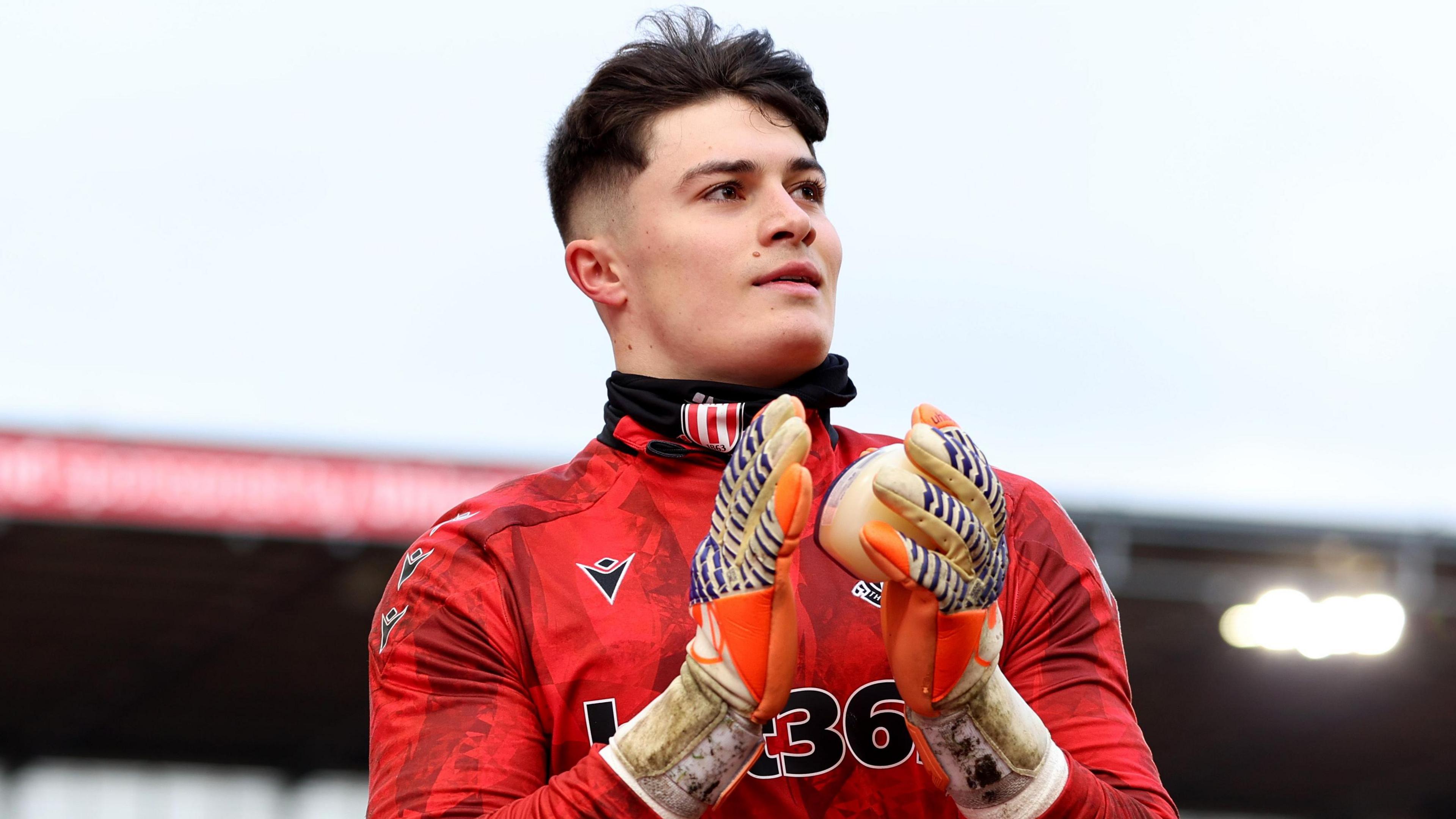 Tommy Simkin, wearing a red Stoke City warm-up top, claps his hands together with goalkeeping gloves on before a game for the Potters.