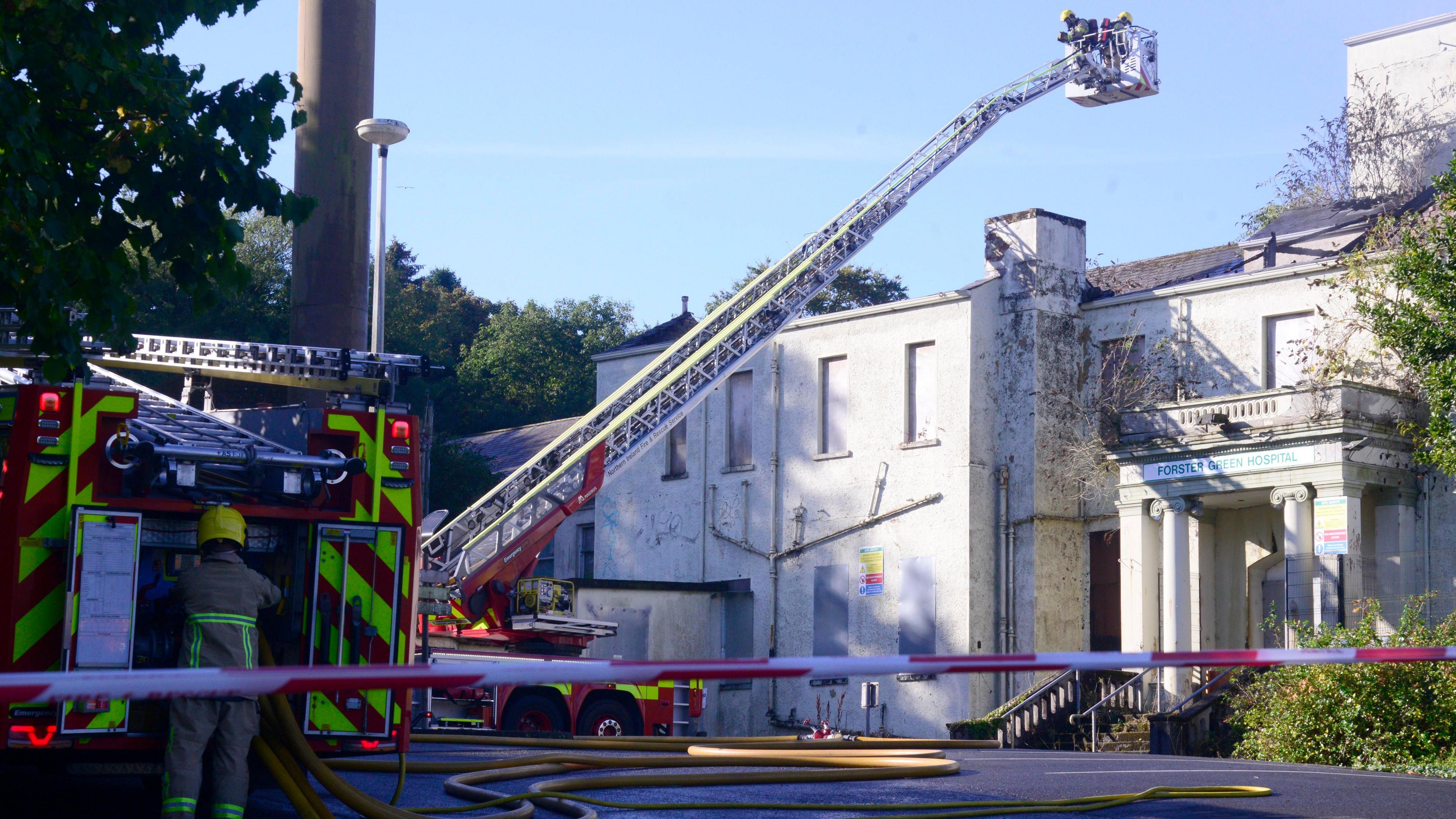 A fire truck is parked beside a white derelict building with a high appliance towering above. One firefighter is standing at the back of the truck while two others are at the top of the appliance, looking down on the building.