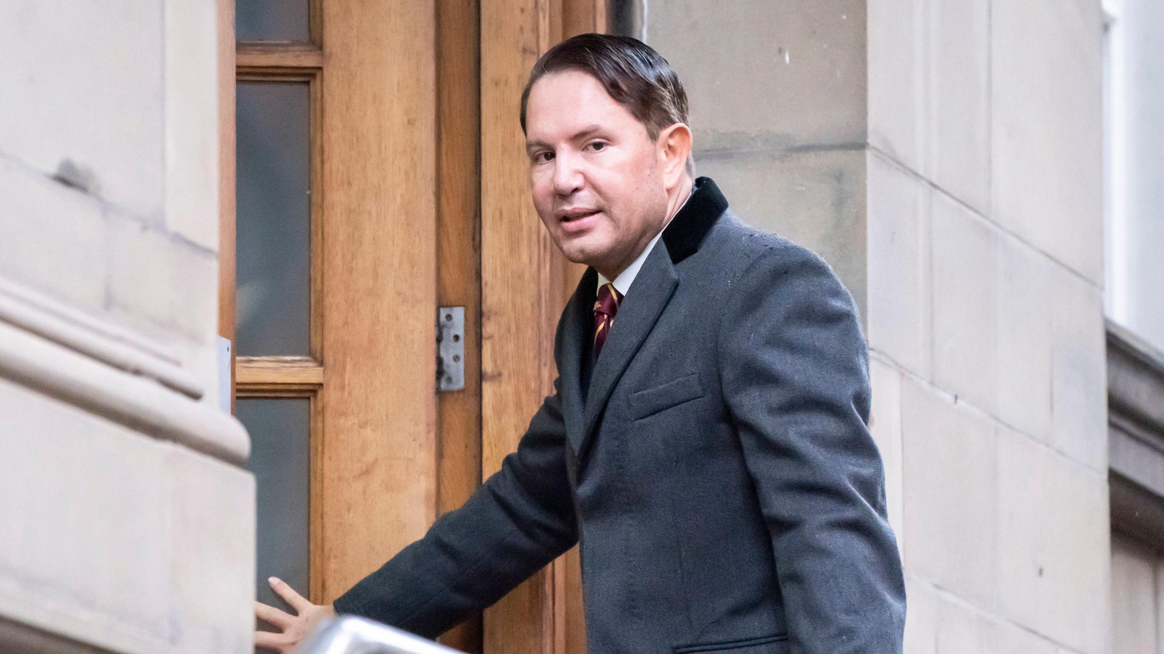 A smartly dressed James Stunt, with dark hair swept to one side, enters a building wearing a grey coat.