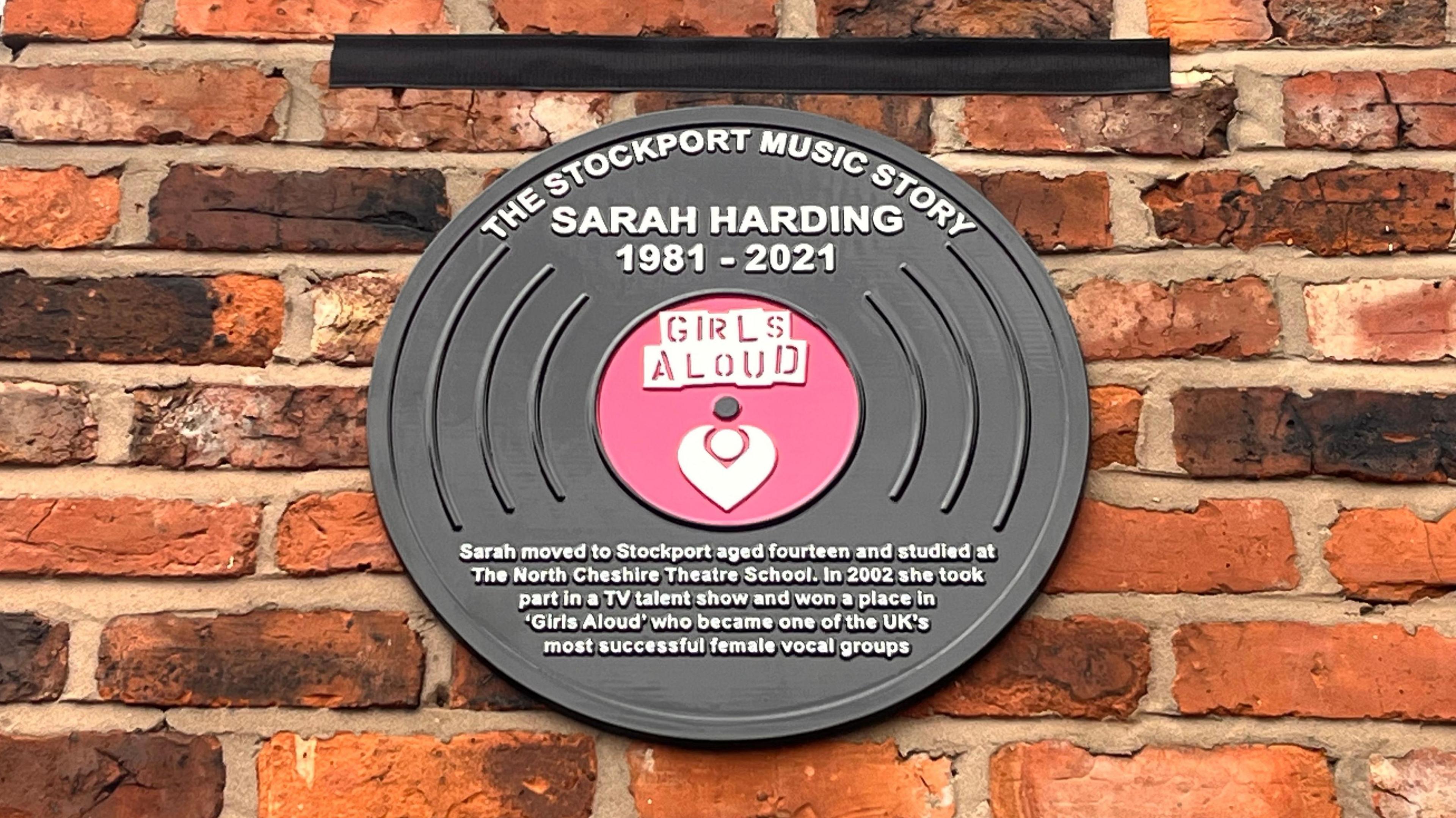 A black and pink plaque styled to look like a vinyl disc on a brick wall, with a short tribute to Sarah Harding and a logo for the band Girls Aloud 