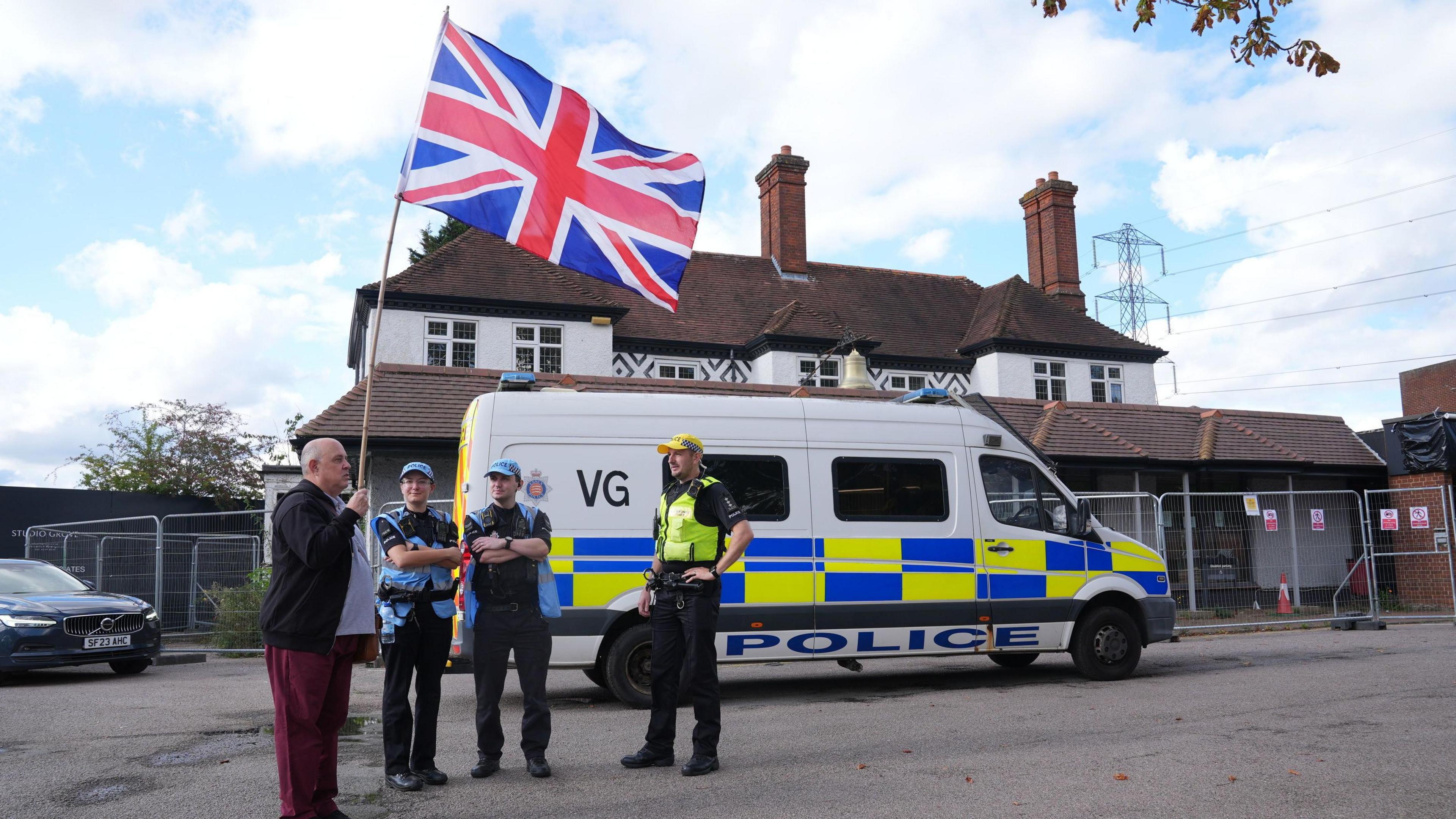 A house which is a hotel with a police van and three police officers standing in front of it with an older man holding a large union jack flag.
