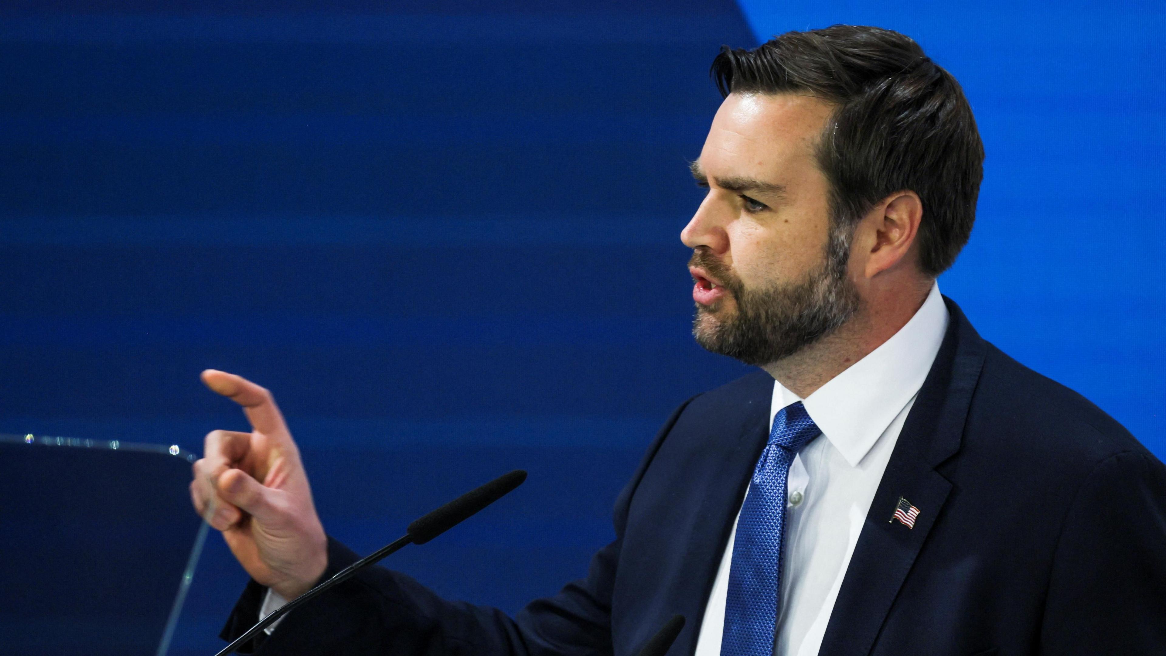 JD Vance, wearing a dark blue suit and blue tie with a US flag lapel badge, points his finger while giving a speech at the Munich Security Conference.