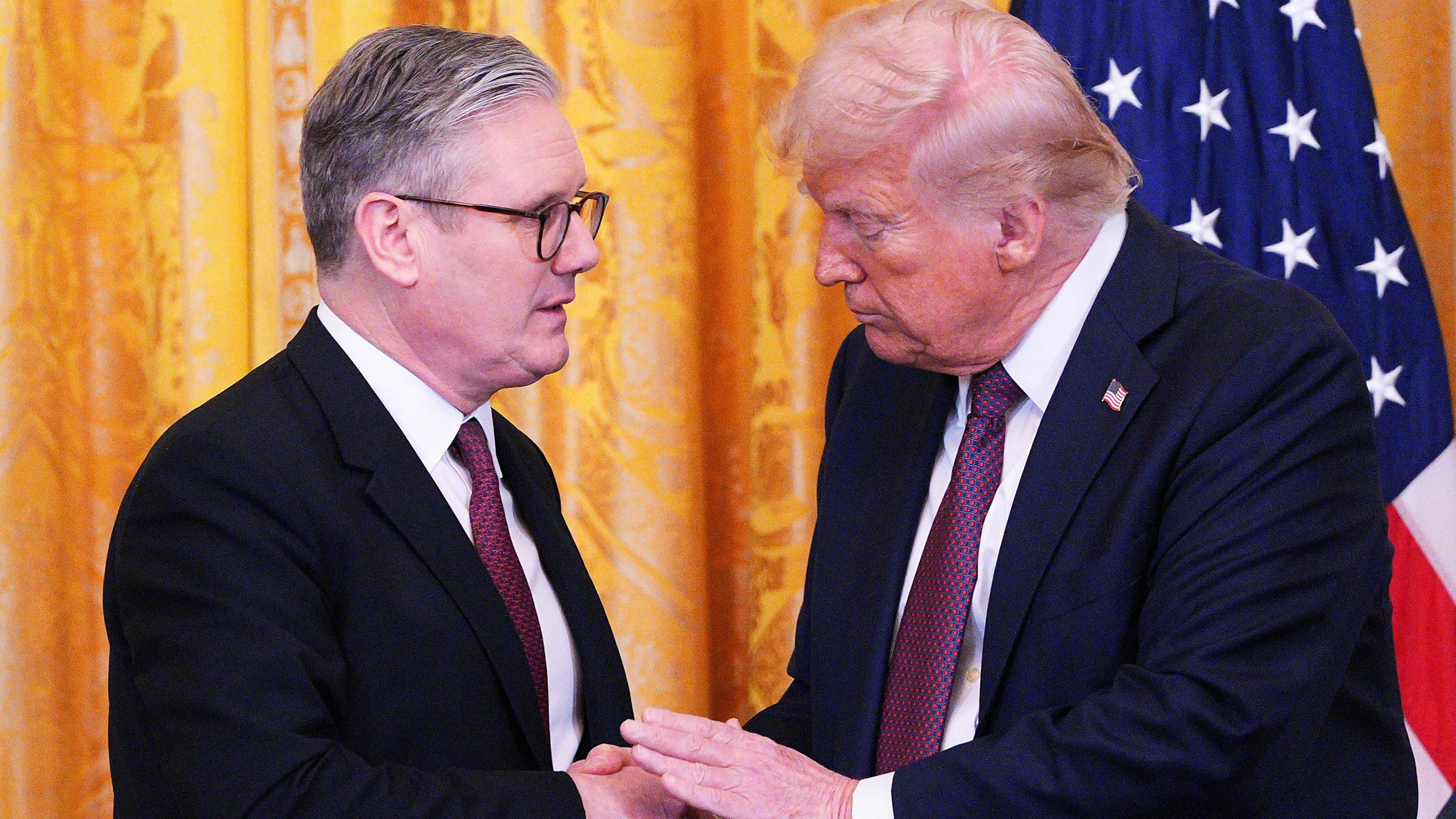 Sir Keir Starmer and Donald Trump, both wearing suits and ties, shake hands, with an orange curtain and US flag in the background