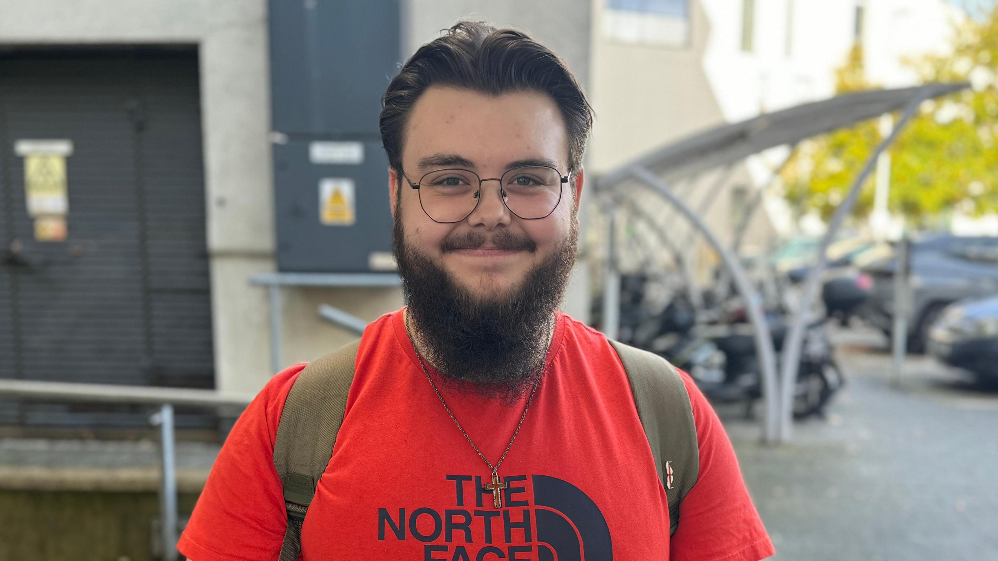 A young man with black hair, glasses and a beard wearing a red t-shirt smiles at the camera