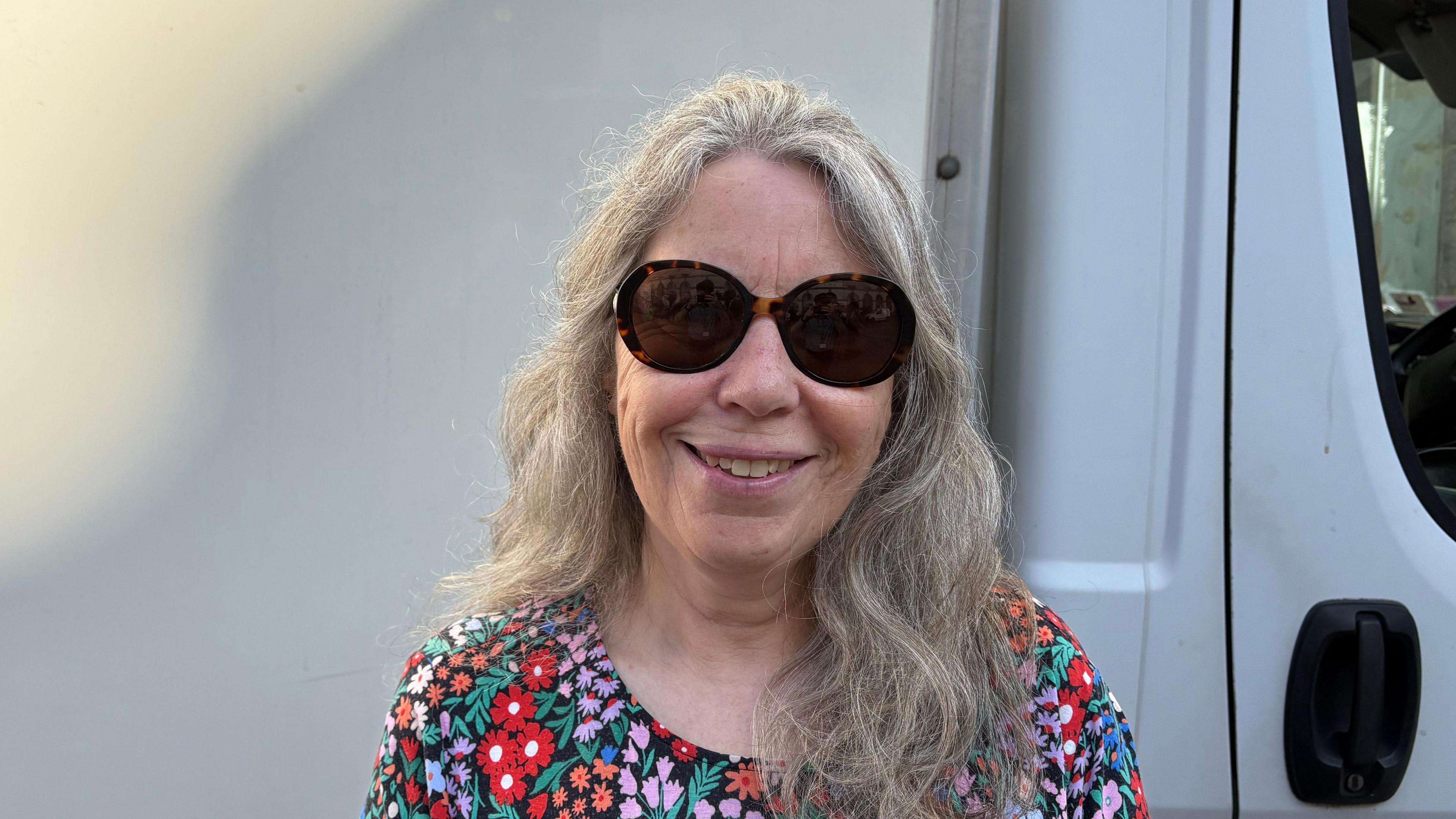 A lady with long grey hair and brown sunglasses looking at a camera. She's standing in front of a big white van.