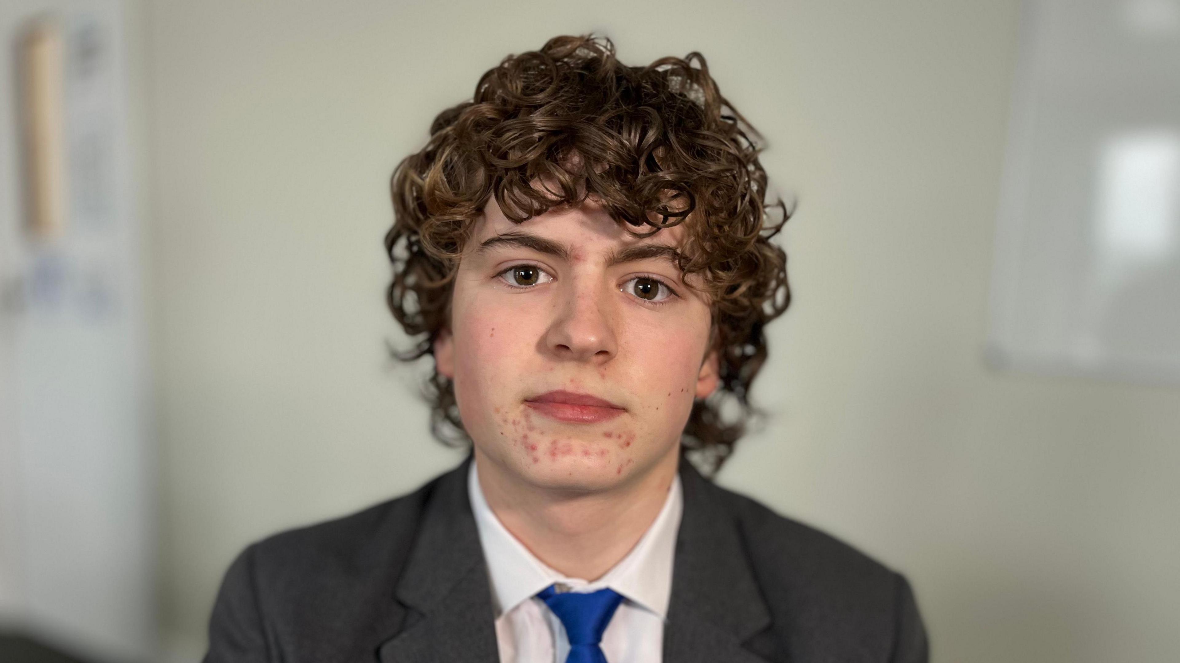 Josh is giving a neutral expression towards the camera. He's wearing a white shirt, blue tie and grey blazer