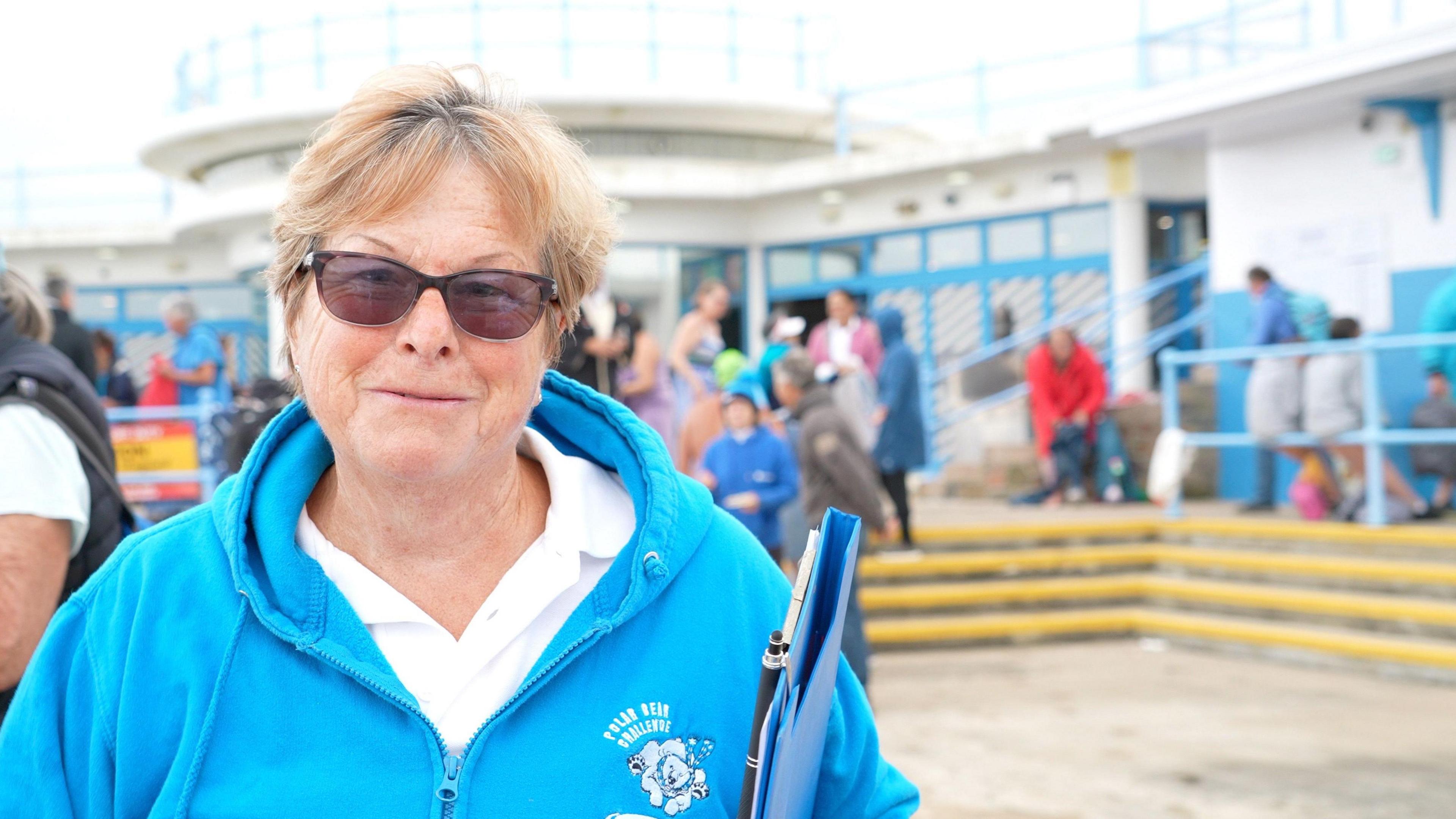 Sally Minty-Gravett wearing a blue hoody and holding a clipboard. She has short brown hair and glasses.