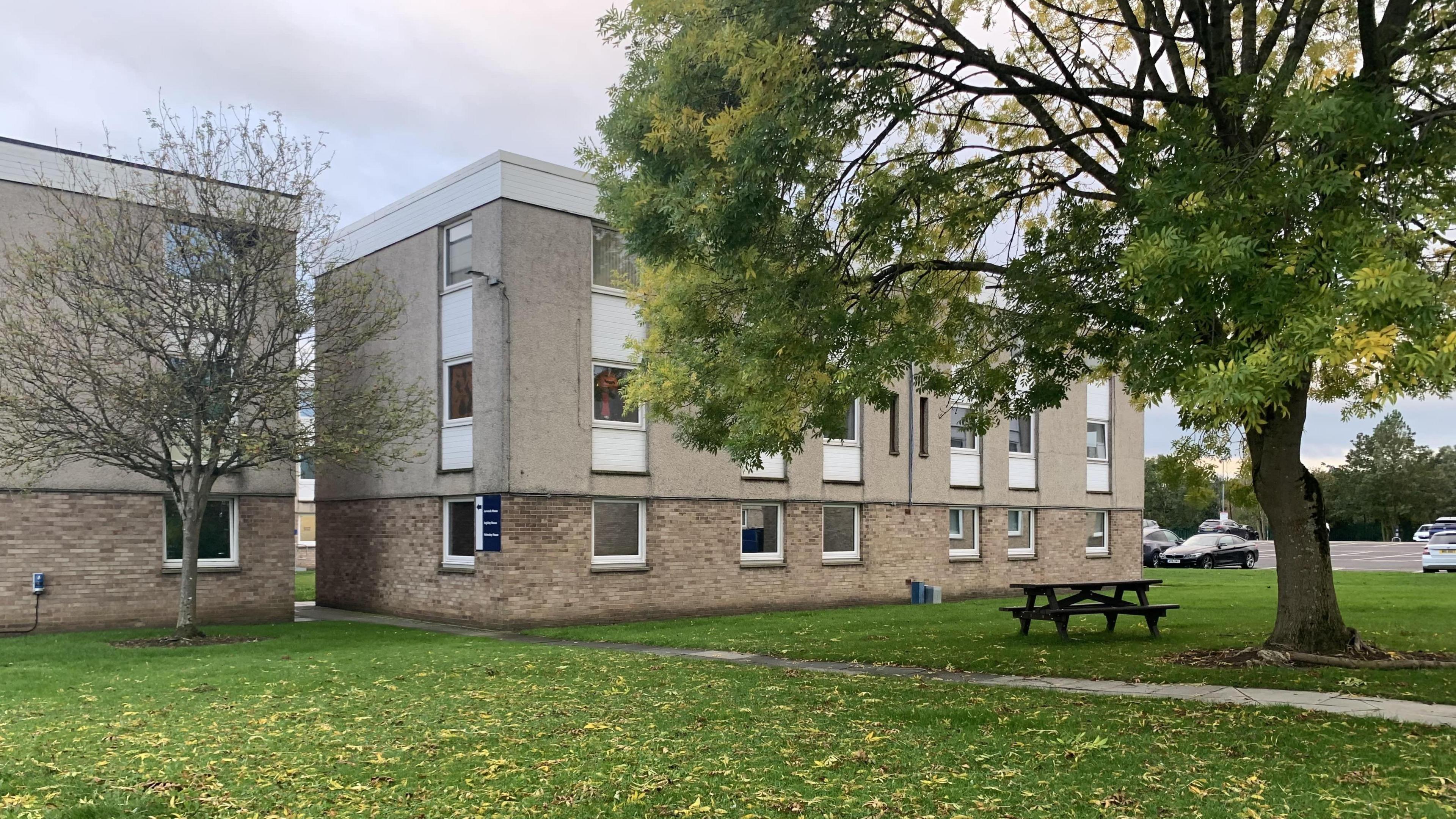 Office blocks at North Tees NHS Trust, standing on a grass lawn with a picnic table and trees and yellow leaves on the grass. There is a car park, with cars, behind them.