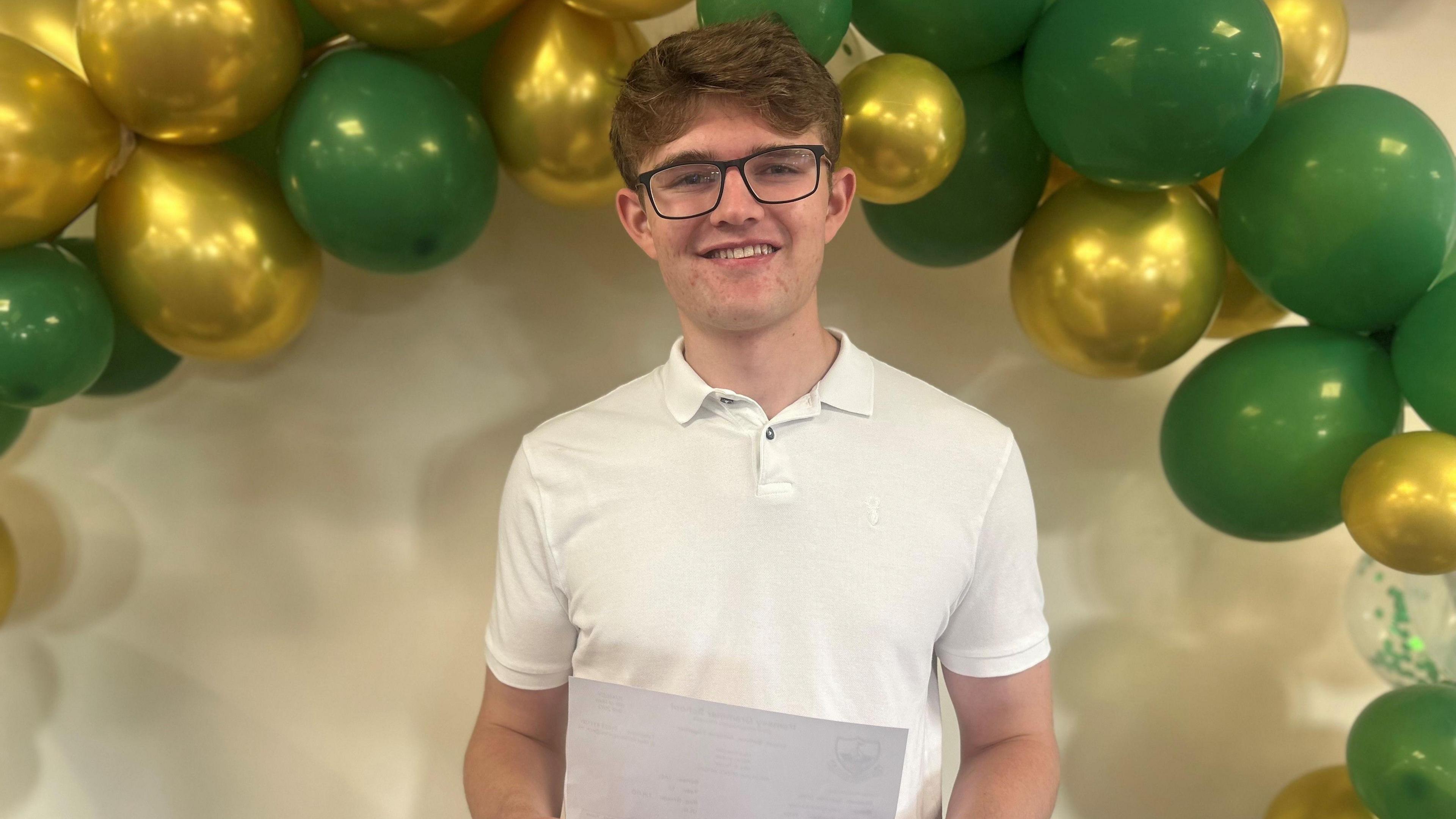 Leo Crowe wears a white shirt and dark spectacles. He holds a piece of paper with his results and stands under an arch with green and gold balloons.