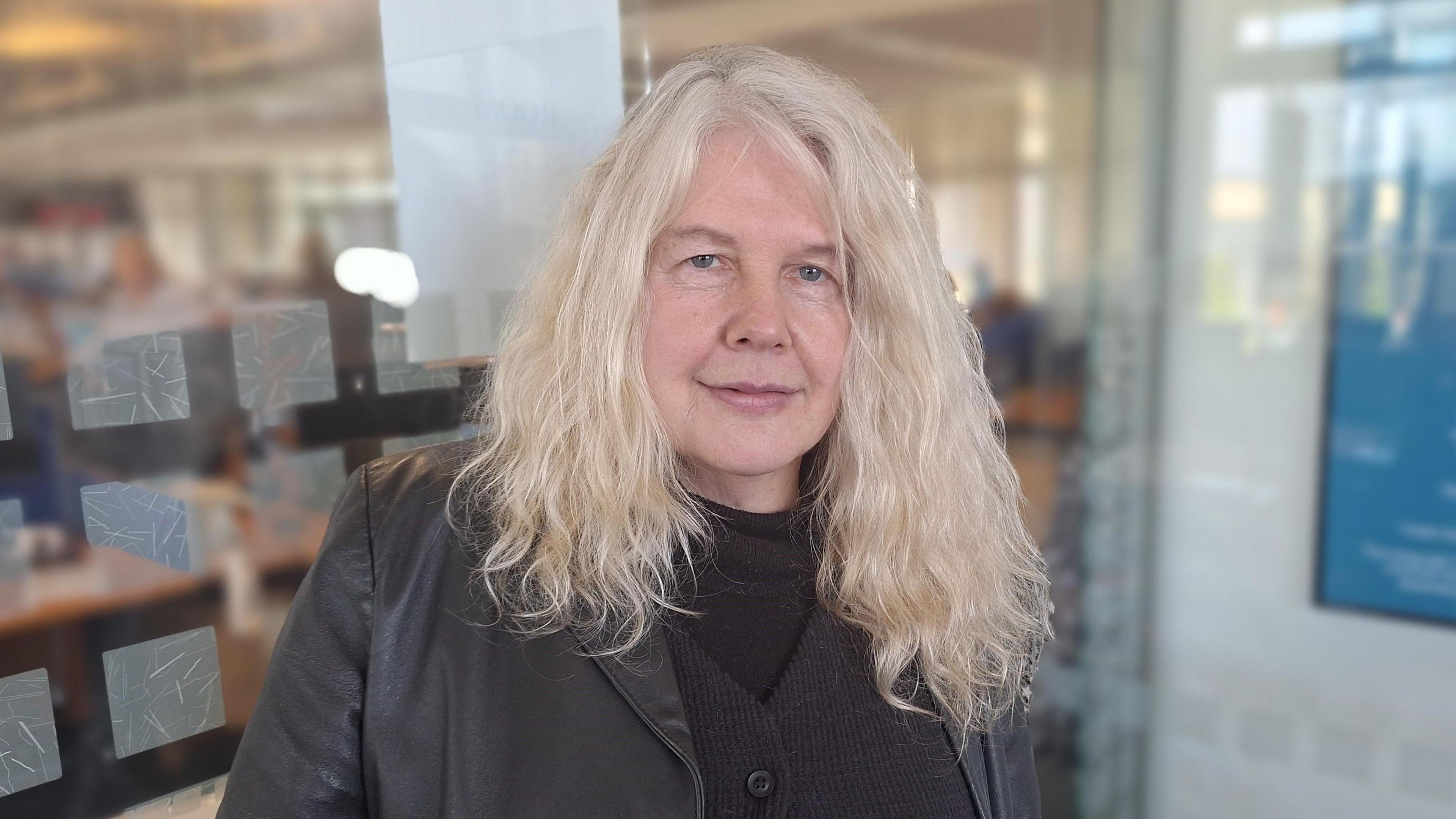 Linda Davies stands in front of a glass office door. She has long blonde hair and wears all black. She is looking into the camera.