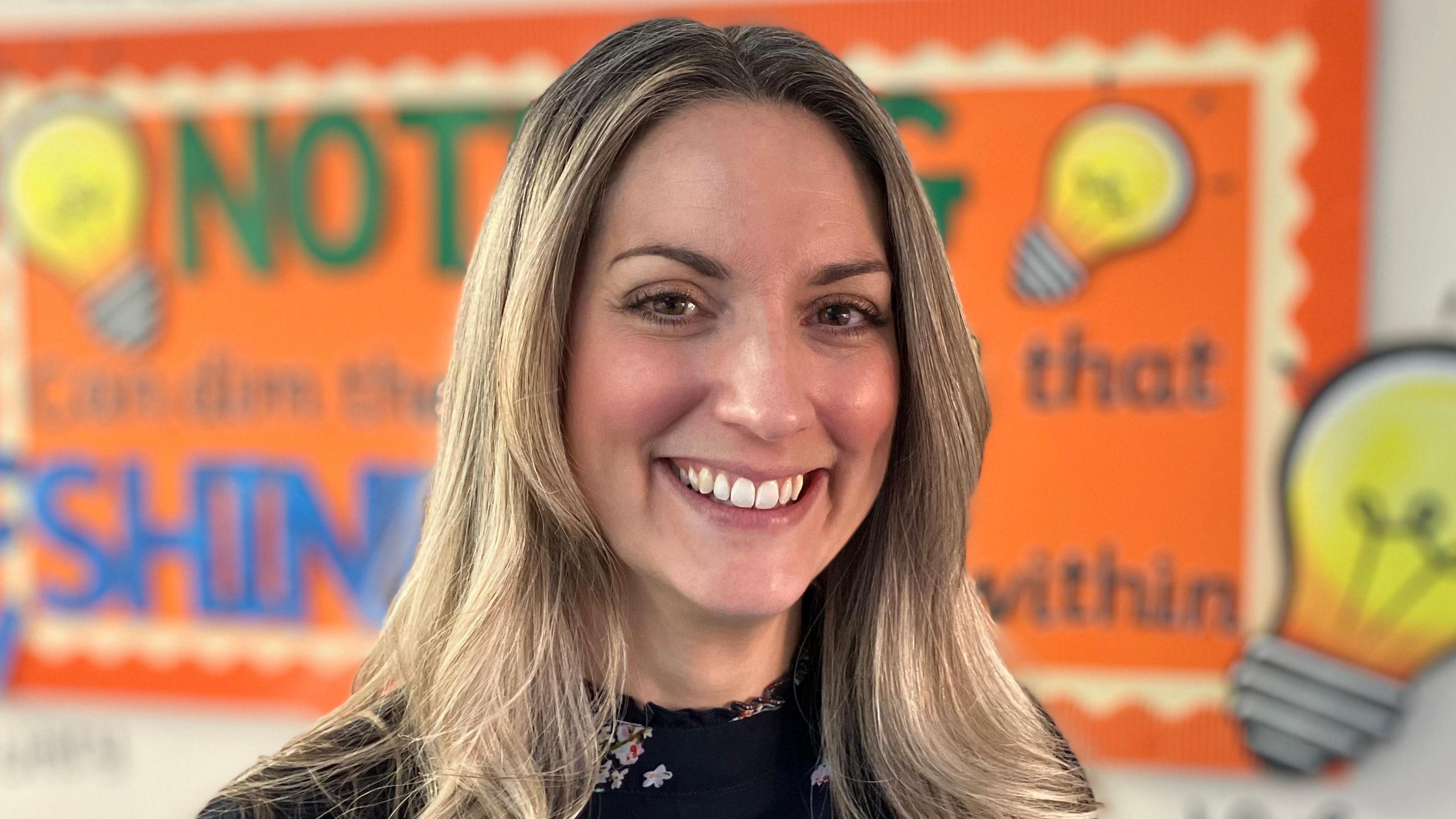 Headteacher Fiona Booth is shown at head and shoulders. She is standing with shoulder-length blonde hair and smiling at the camera, in front of a colourful school display board. 