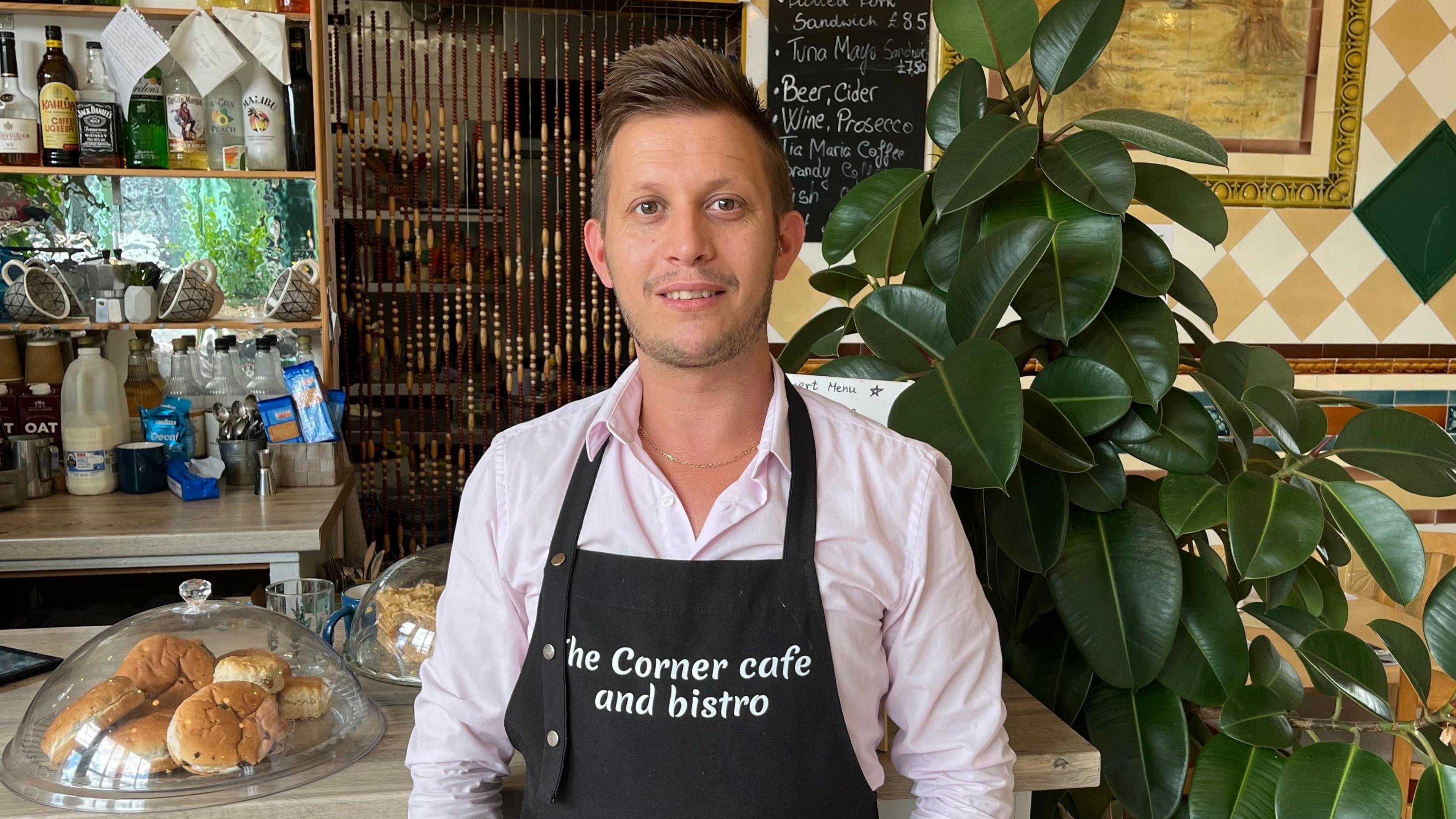 A man stands behind the counter of a café wearing a pink shirt and a black apron with the words "The Corner cafe and bistro". The counter holds containers of buns and cakes. Bottles containing alcohol are visible on shelves and a chalkboard menu is also hanging on the wall. A green plant and decorative tiles are also visible.